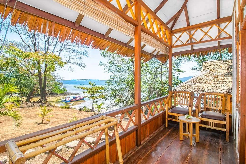 A balcony in a diving resort cabin with woods, ratans walls and furnitures combined, and a roof made of bamboo leaves, facing the sea.