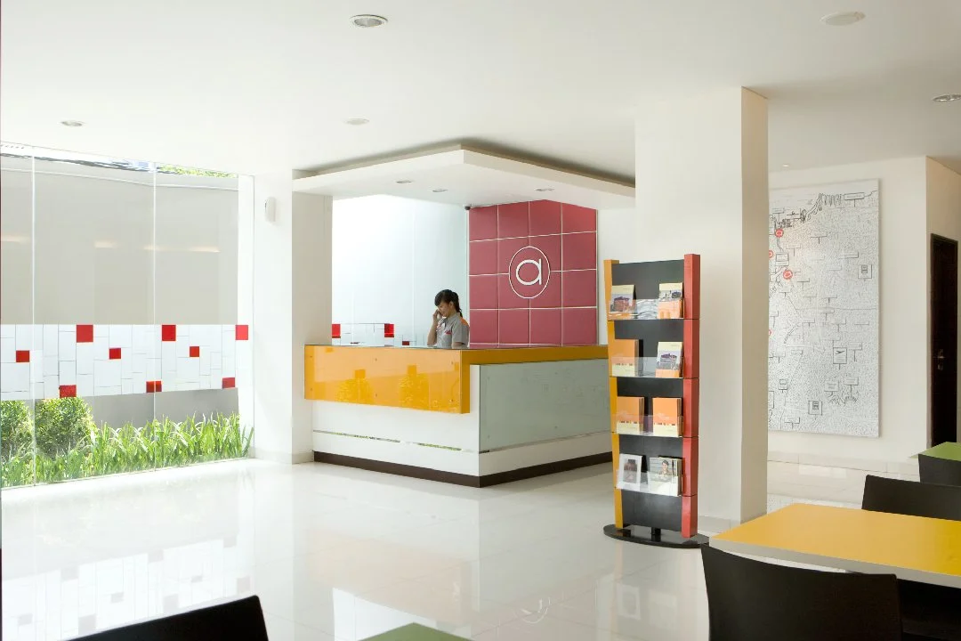 a hotel lobby with a receptionist, plants outside, leaflets and books for reading, tables and chairs.