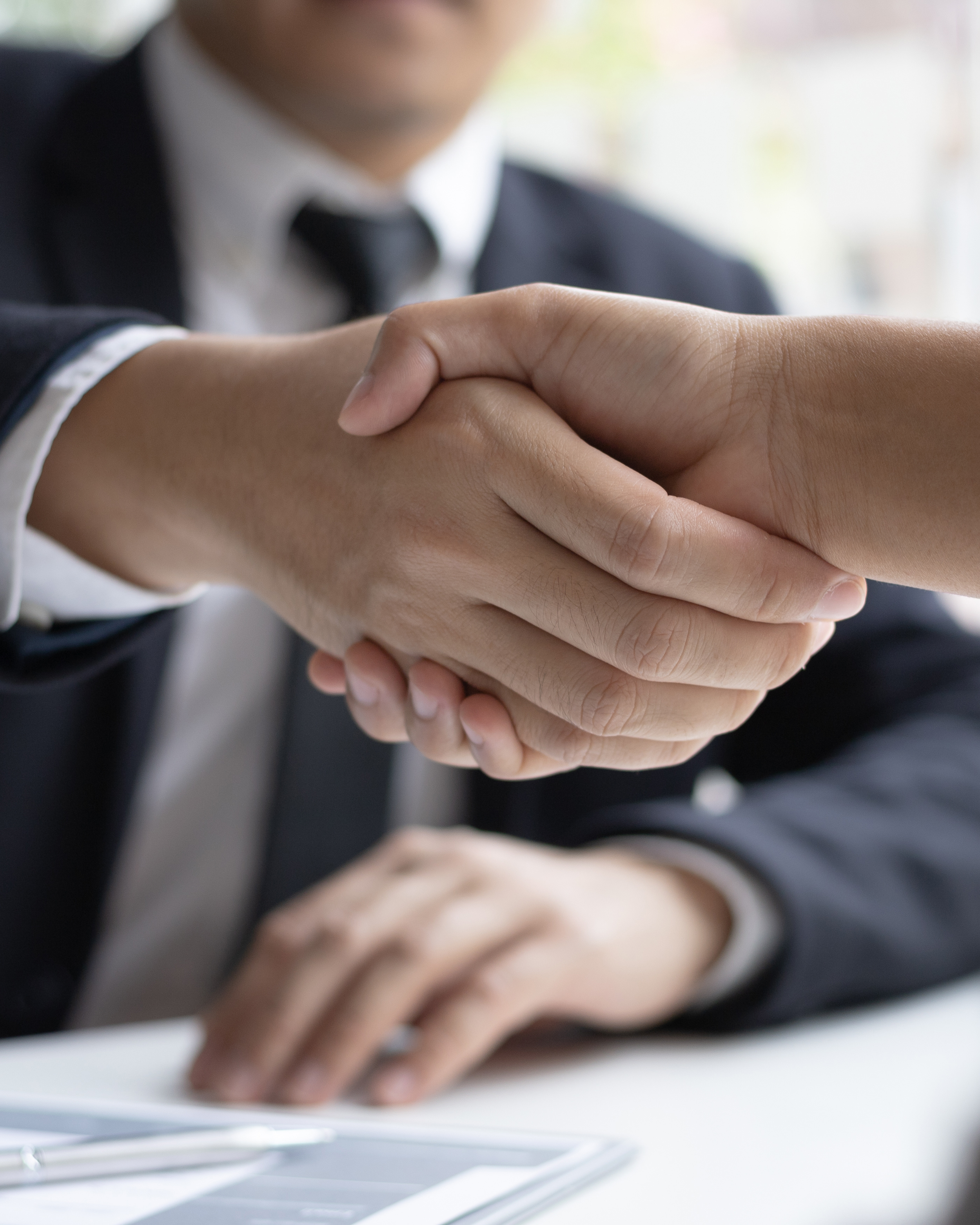 Close up of two hands clasped together on a table, with a person in a black suit and white shirt in the background.