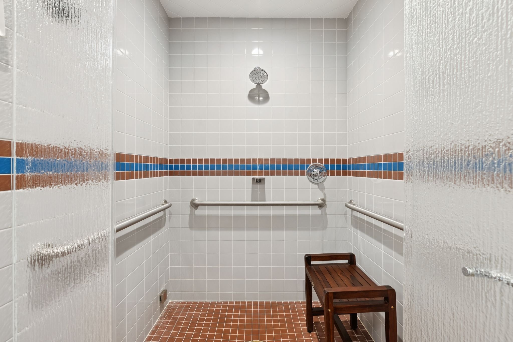A walk-in shower with white tiled walls, a horizontal row of brown and blue tiles, a silver showerhead, and a small wooden bench inside.