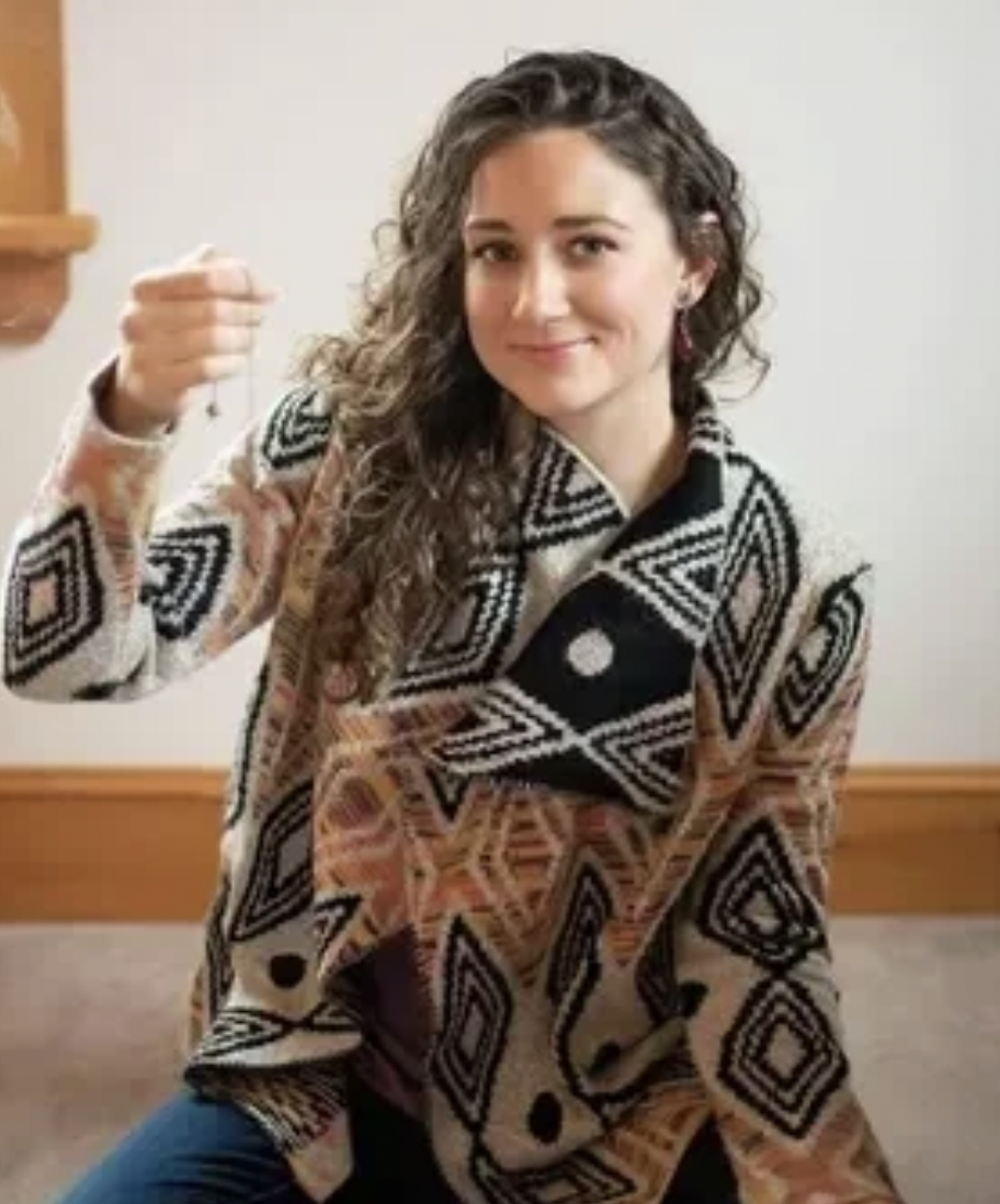 A young woman with curly hair sitting indoors, wearing a patterned sweater and holding a necklace.