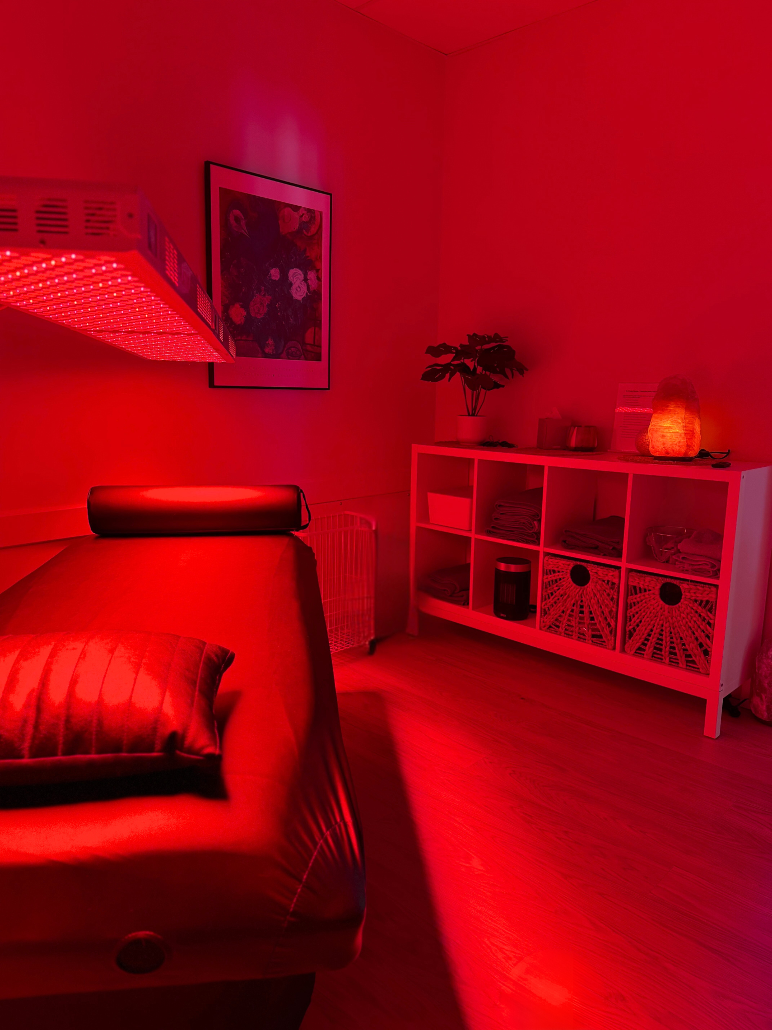 A room illuminated with red lighting. There is a black couch with a ribbed cushion, a white shelving unit with folded towels, a potted plant, a salt lamp, and framed artwork on the wall.