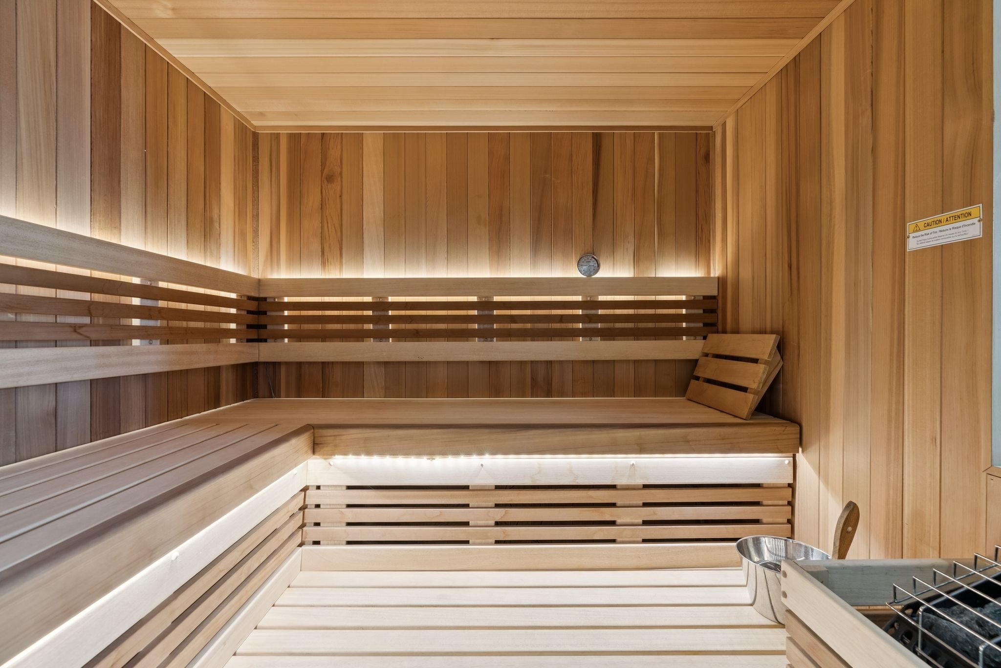 Interior of a wooden sauna with benches, a small round thermometer on the wall, and a metal heater in the corner.