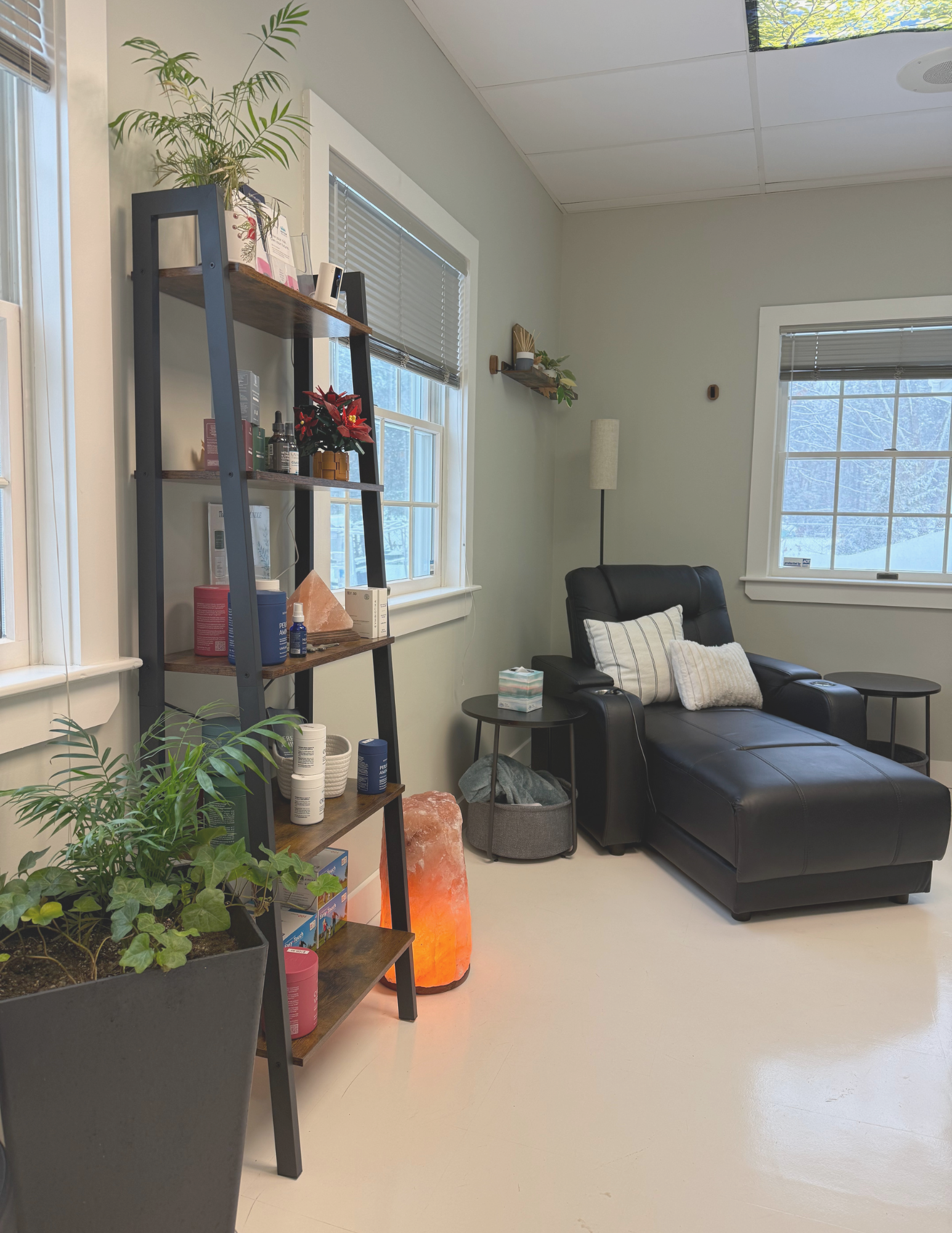 A cozy corner with a black leather sofa, pillows, a side table, a salt lamp, plants, and windows showing a snowy outdoor scene.