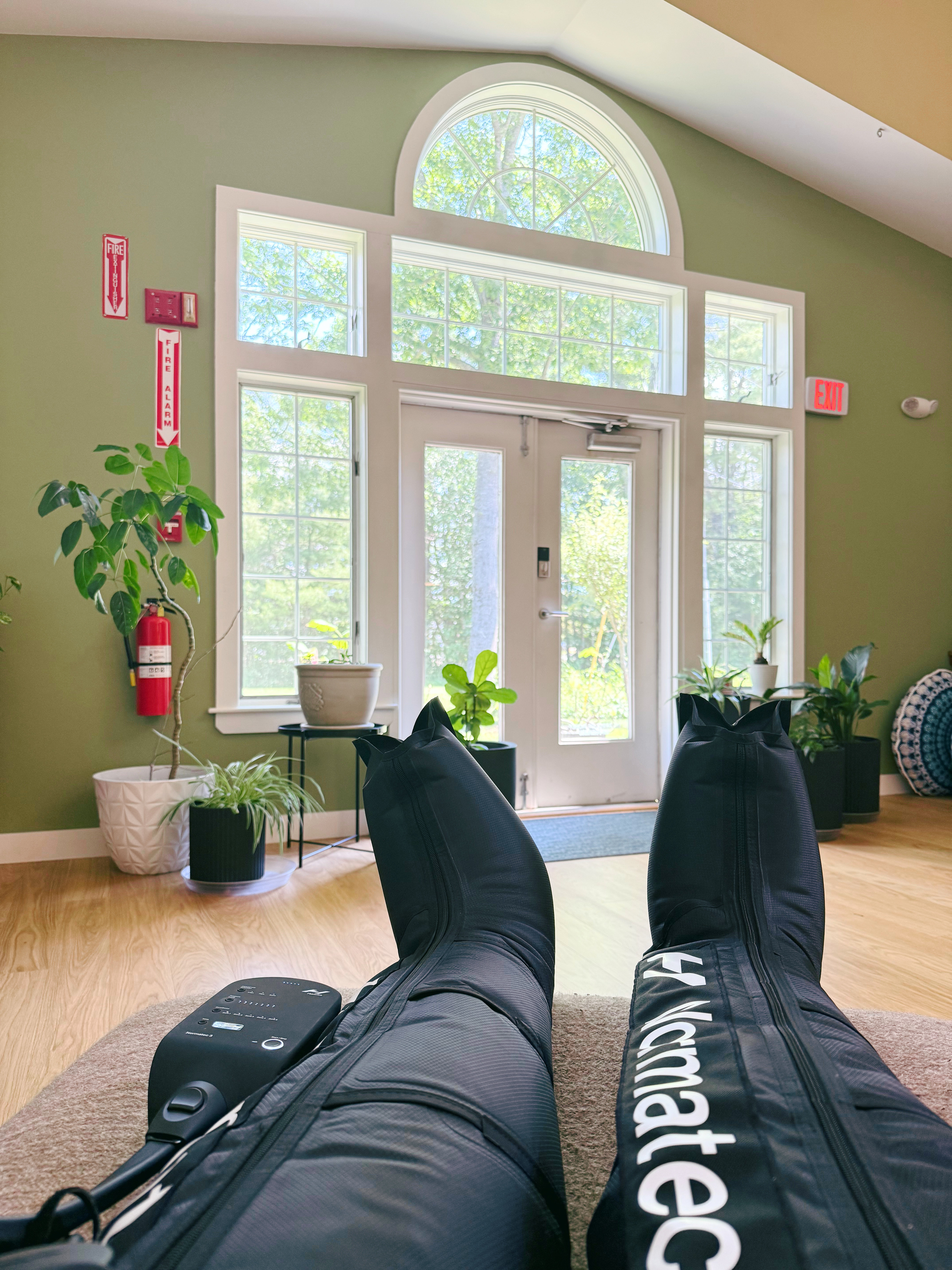 Person lying on their back with legs stretched out, wearing black athletic pants with white lettering, and black shoes, in a room with green walls, large windows, and potted plants visible outside and inside.