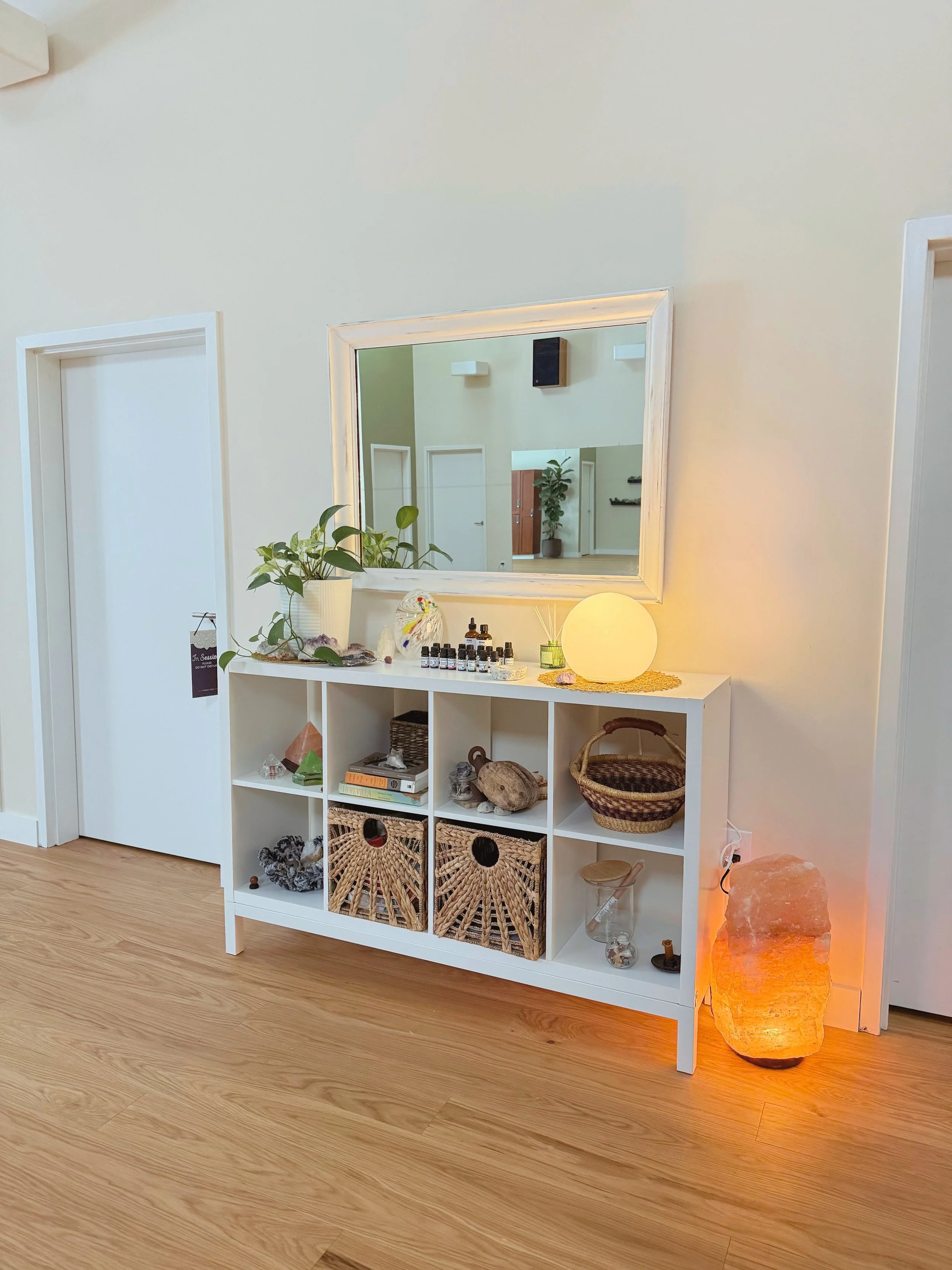 White hallway console table with decorative items including a potted plant, small bottles, candles, and woven baskets, positioned against a light-colored wall with a large mirror and a salt lamp on the right, and hardwood flooring.