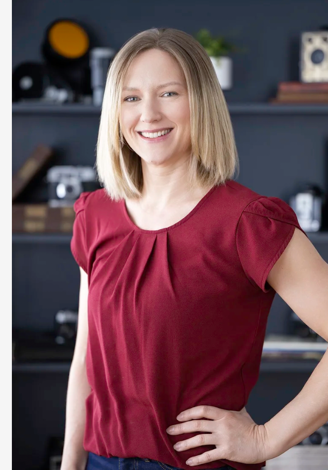 A woman with shoulder-length blonde hair smiling in a red top with a pleated front, standing indoors with a dark bookshelf in the background.