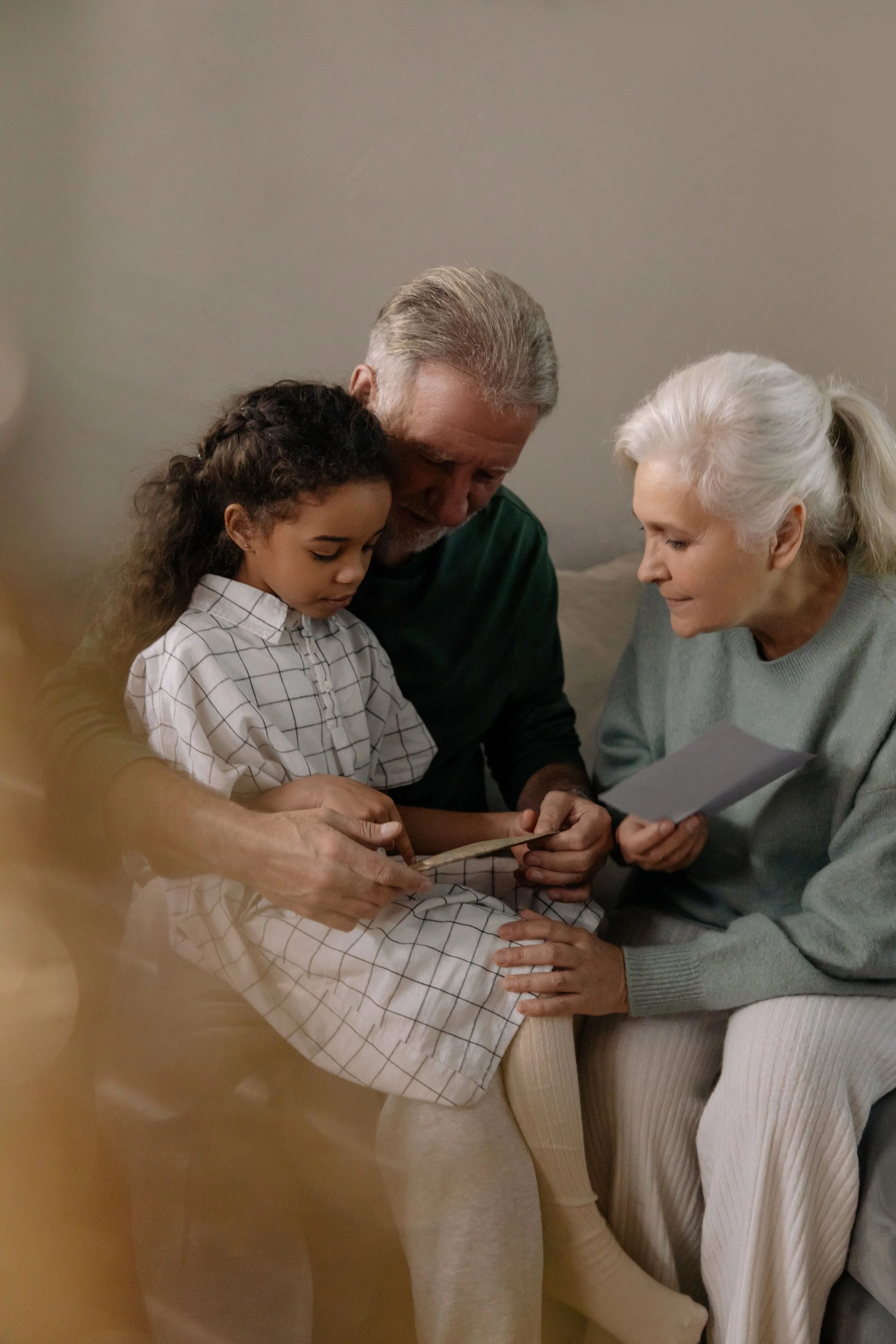 An elderly woman, a middle-aged man, and a young girl are sitting together, looking at some photographs or papers and sharing a moment of connection.