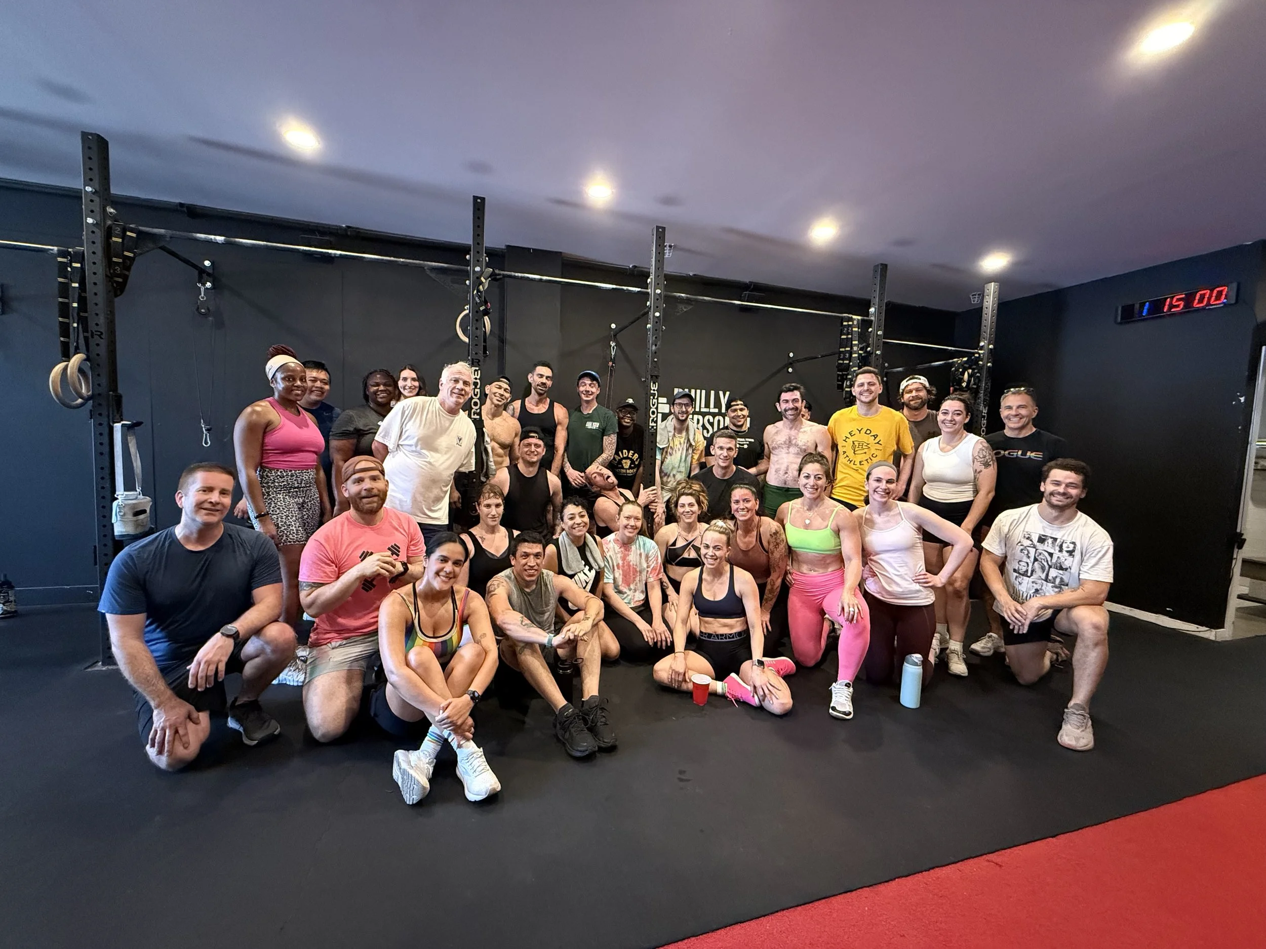 A large group of people gathered in a gym, smiling at the camera, many in workout attire, with gym equipment and a digital clock in the background.