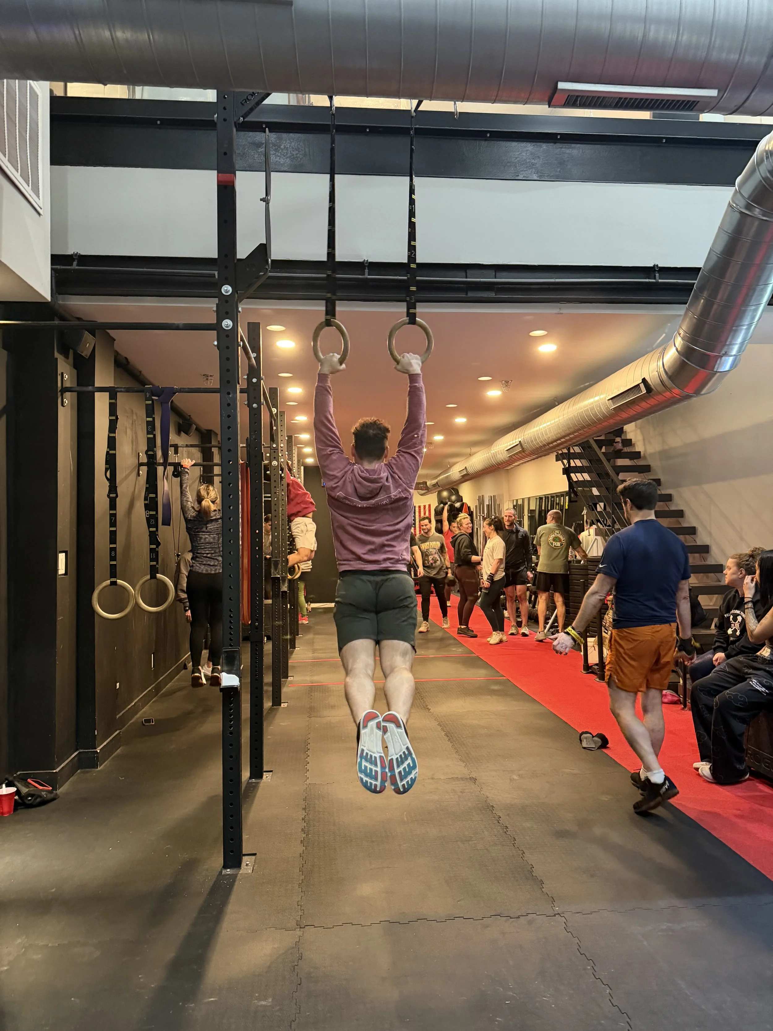 Man in pink hoodie performing a pull-up exercise with gymnastics rings in a gym, surrounded by other people and gym equipment.