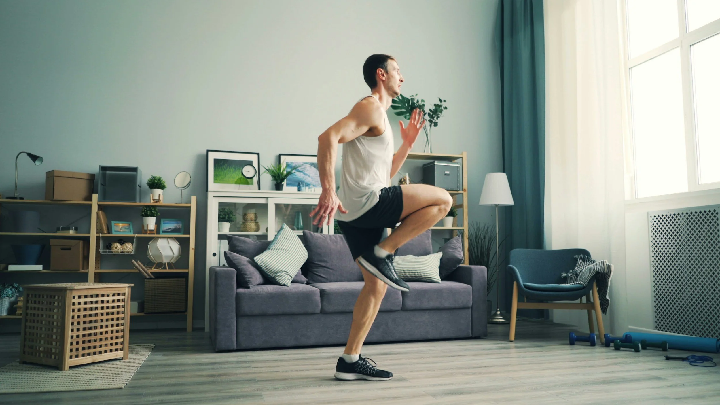 A man is exercising indoors, balancing on a couch with one leg raised and hands in a running position.