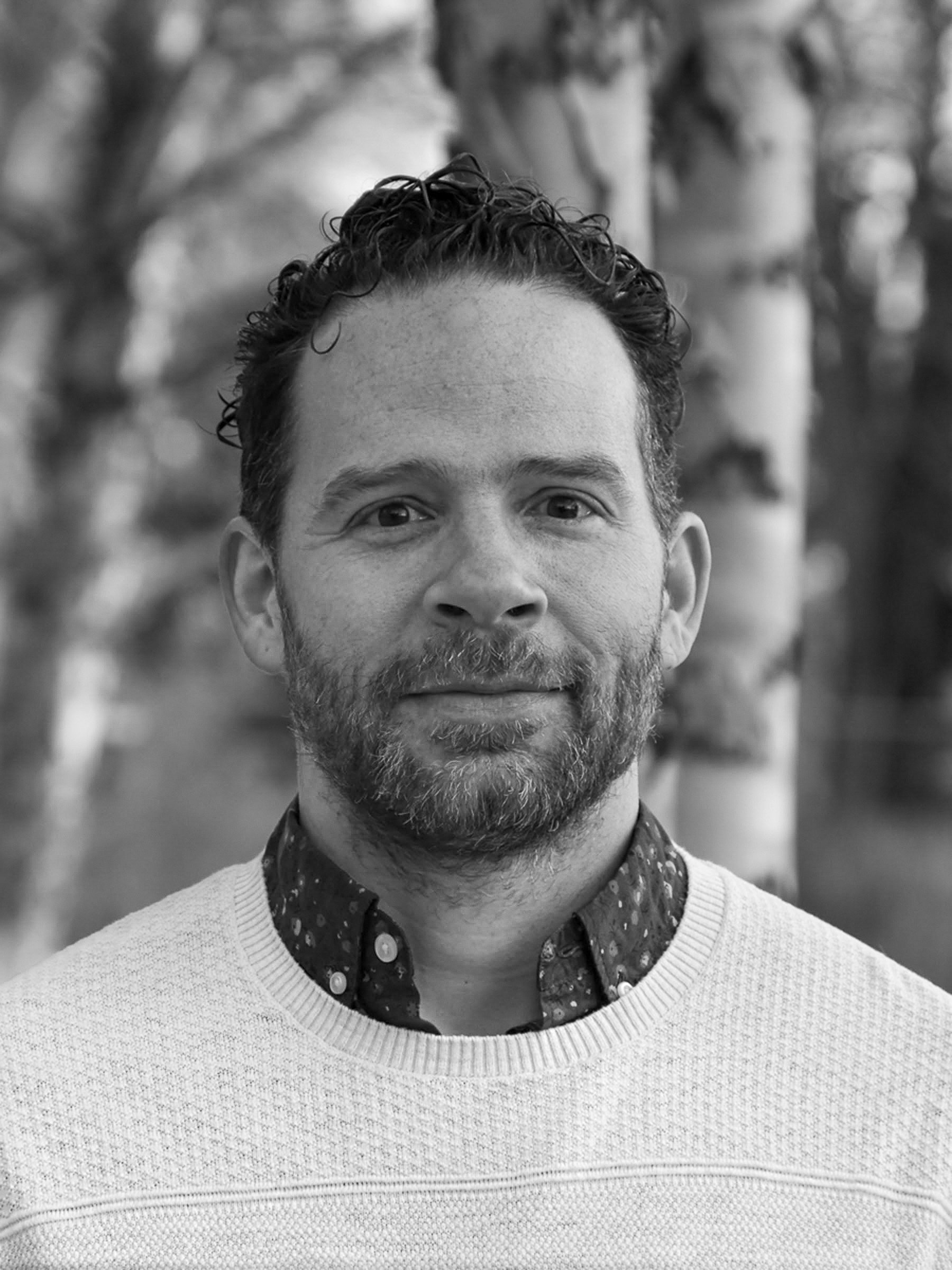 A black and white portrait of a man with short, curly hair and a beard, wearing a sweater over a collared shirt, outdoors with blurred trees in the background.