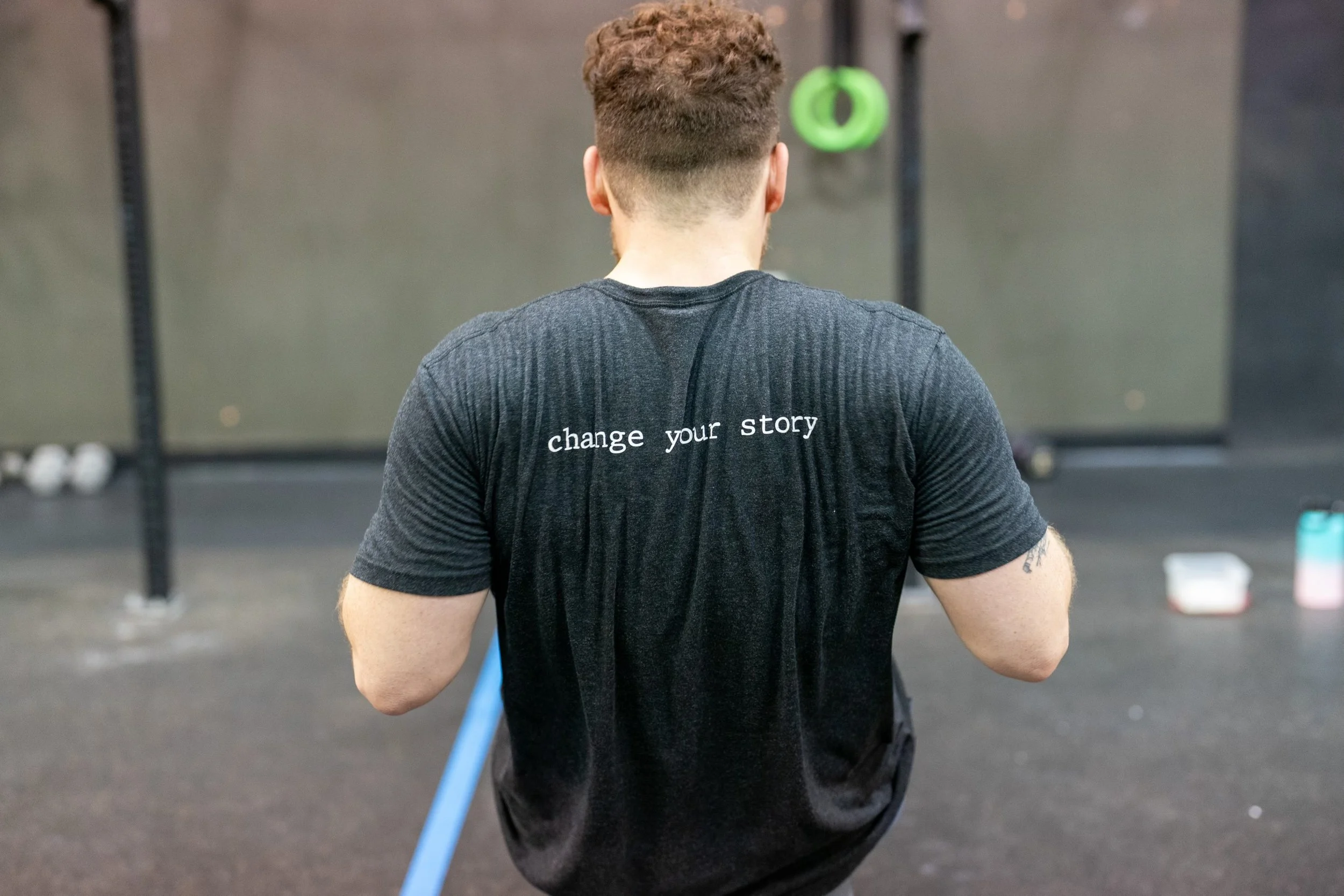 Back of a man with short, curly hair wearing a dark gray t-shirt with the phrase 'change your story' printed on the back, standing in a gym or fitness studio.