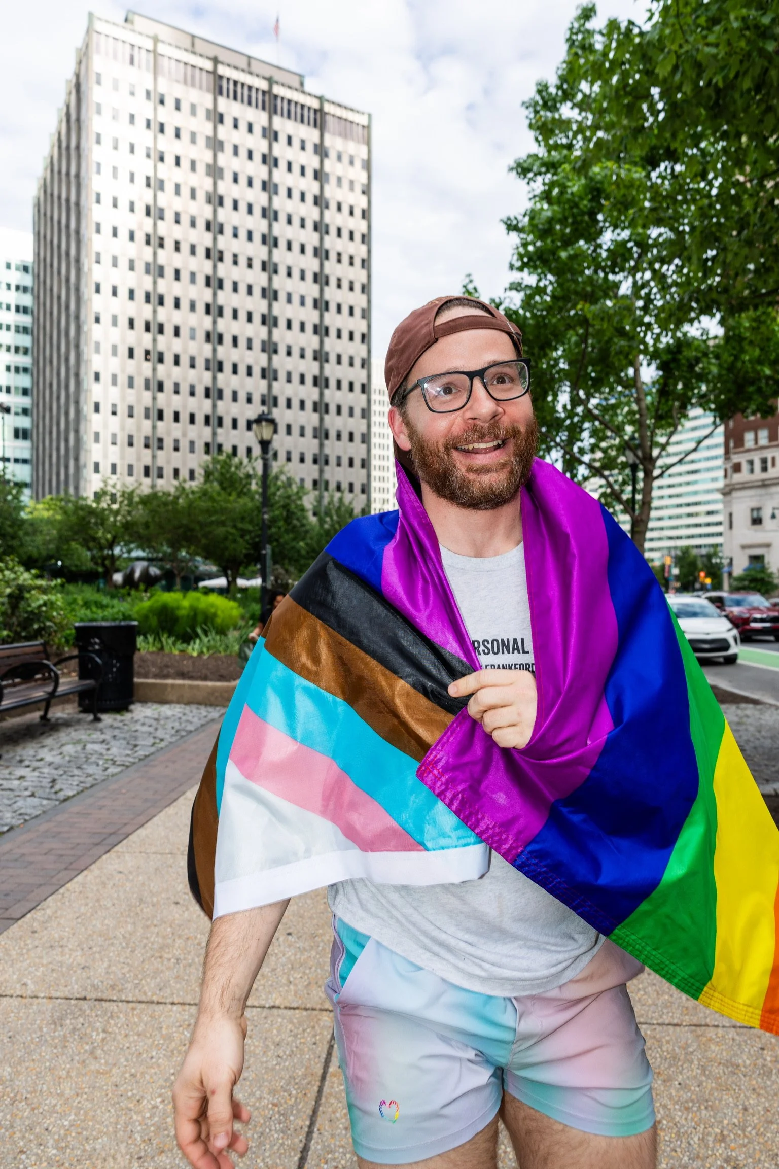 A man with glasses, a beard, and a backward cap smiling and holding a rainbow pride flag in an urban park with tall buildings and green trees.