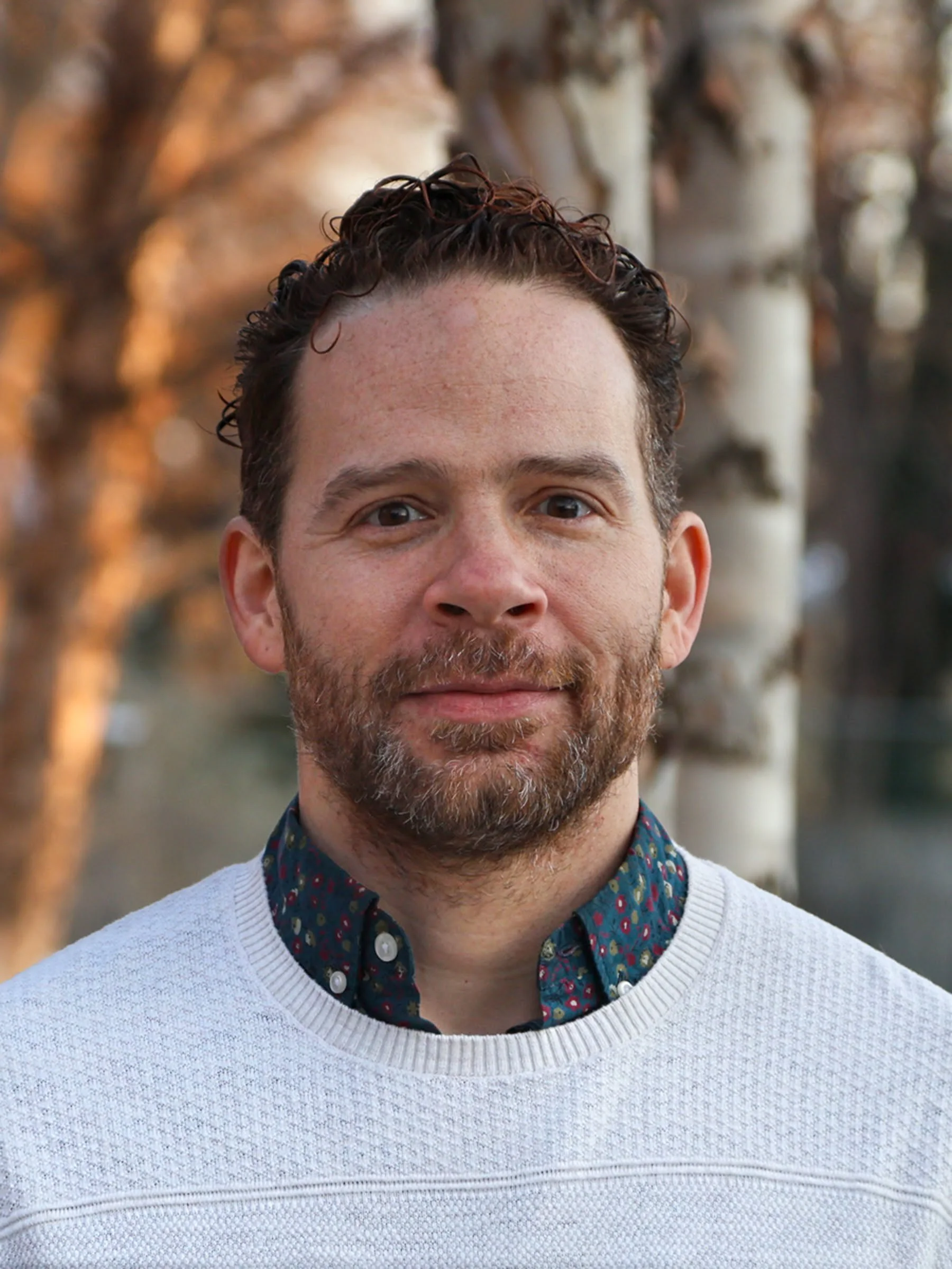 Close-up of a man with short, curly hair and a beard, wearing a white textured sweater over a colorful collared shirt, standing outdoors with blurred autumn trees in the background.
