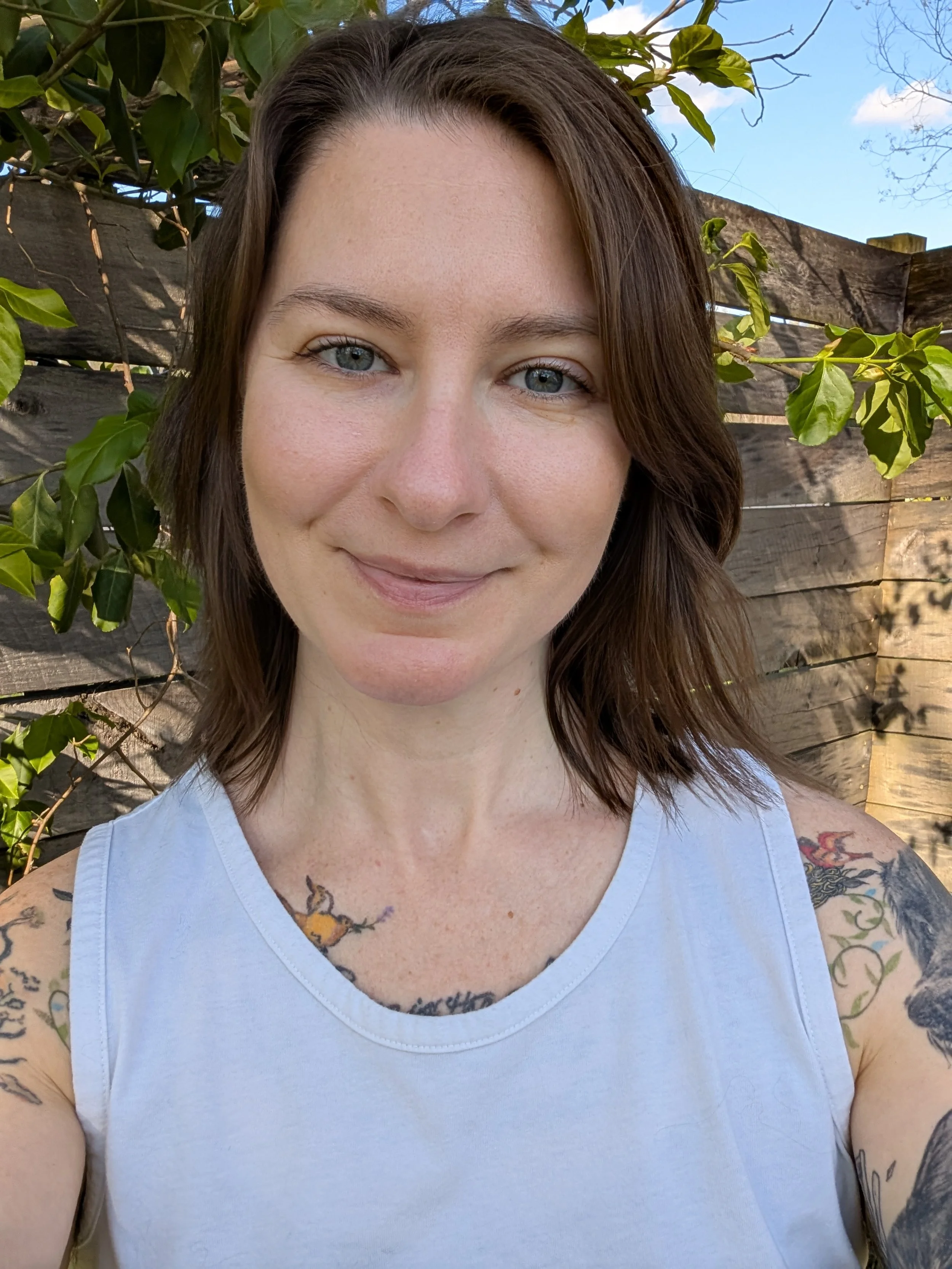 Close-up of a smiling person with tattoos, wearing a white tank top, outdoors near a wooden fence and greenery, with a blue sky in the background.