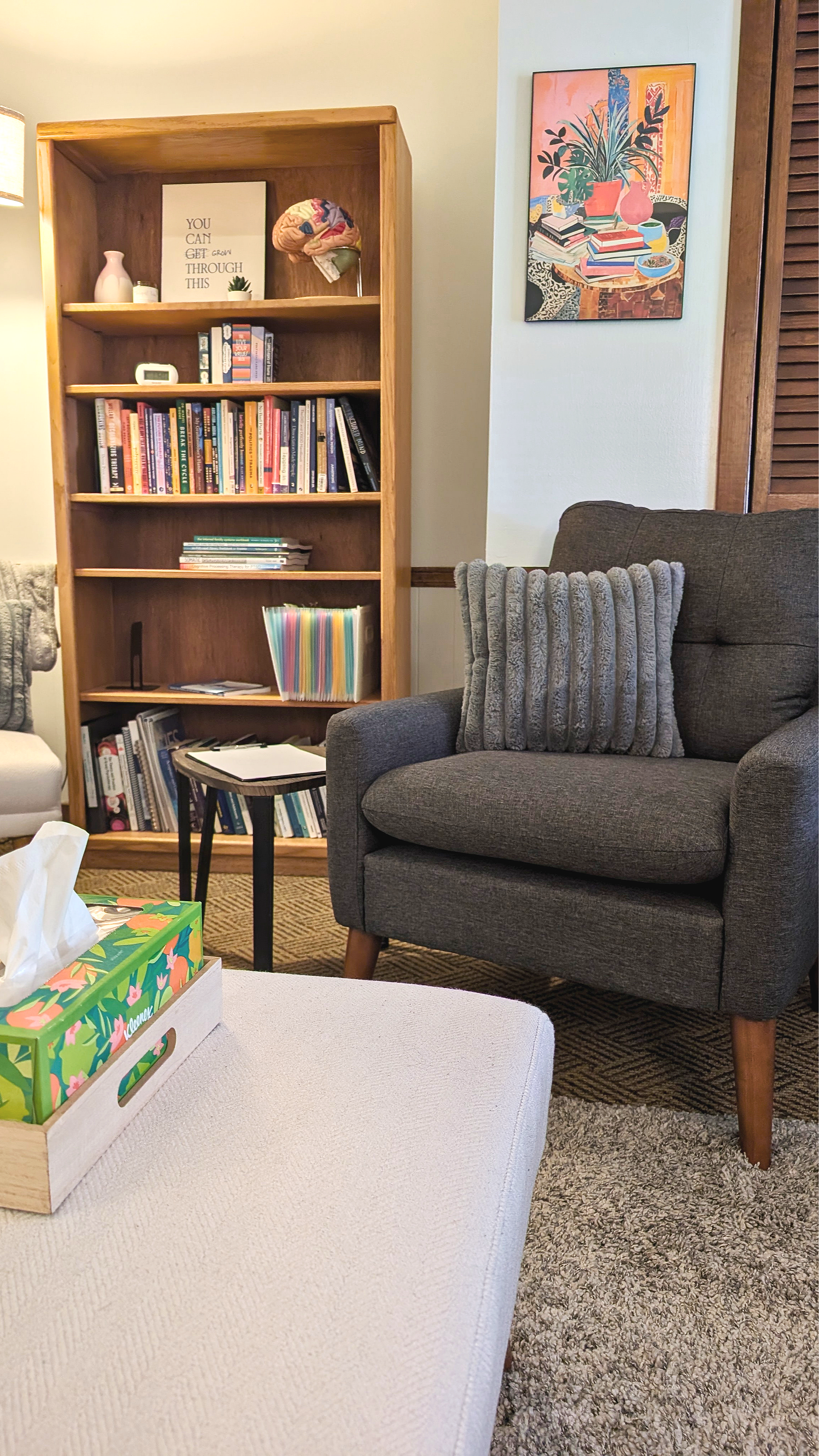 Therapy office with a dark gray armchair and a gray textured pillow, a small black side table with papers, a wooden bookshelf filled with books and decorative items, framed artwork on the wall, and a tissue box on a white ottoman in the foreground.