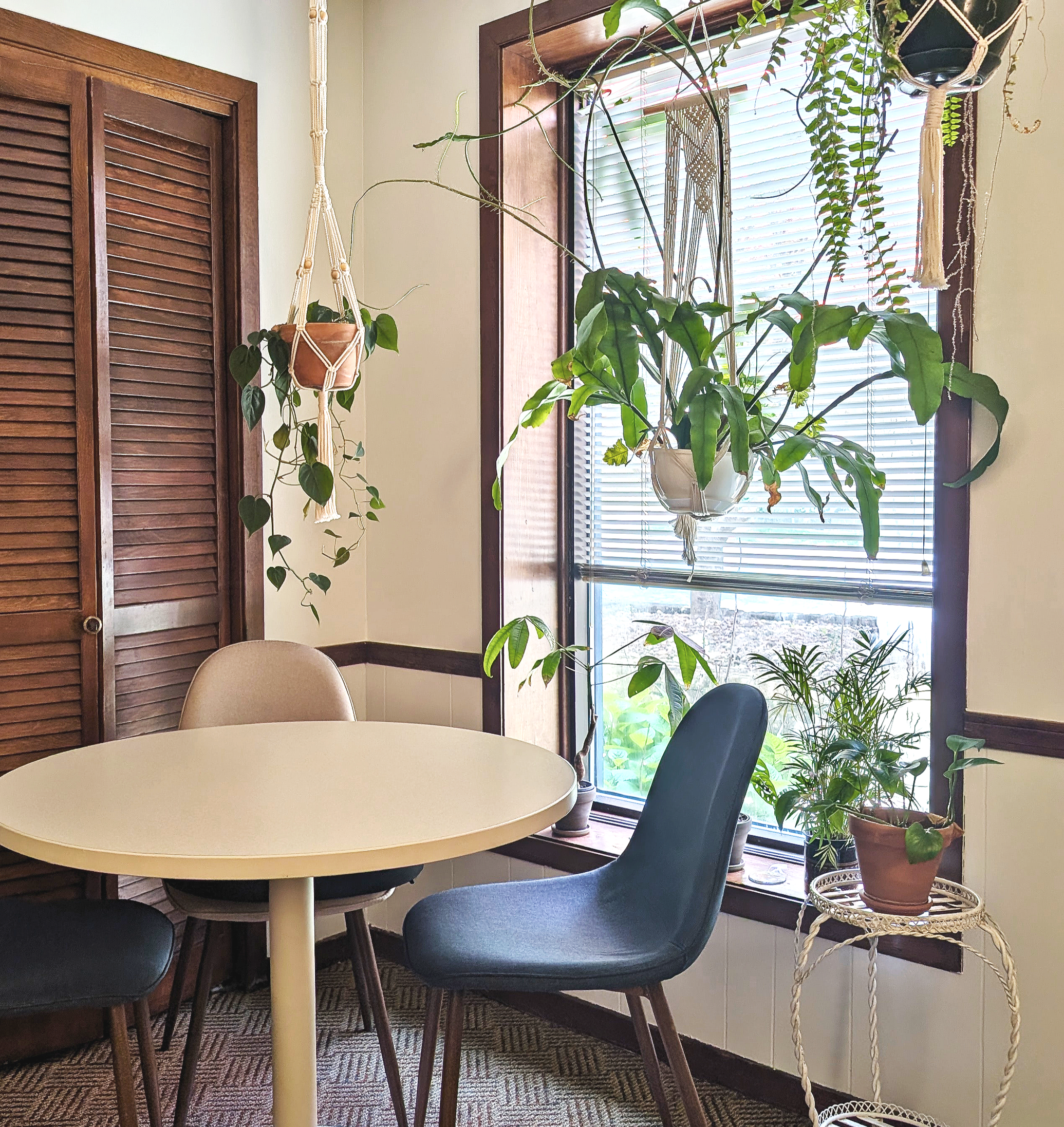 A circular cream-colored table and four chairs sit in front of a window. There are various potted plants, some hanging in macrame planters. The window has wooden framing with blinds.