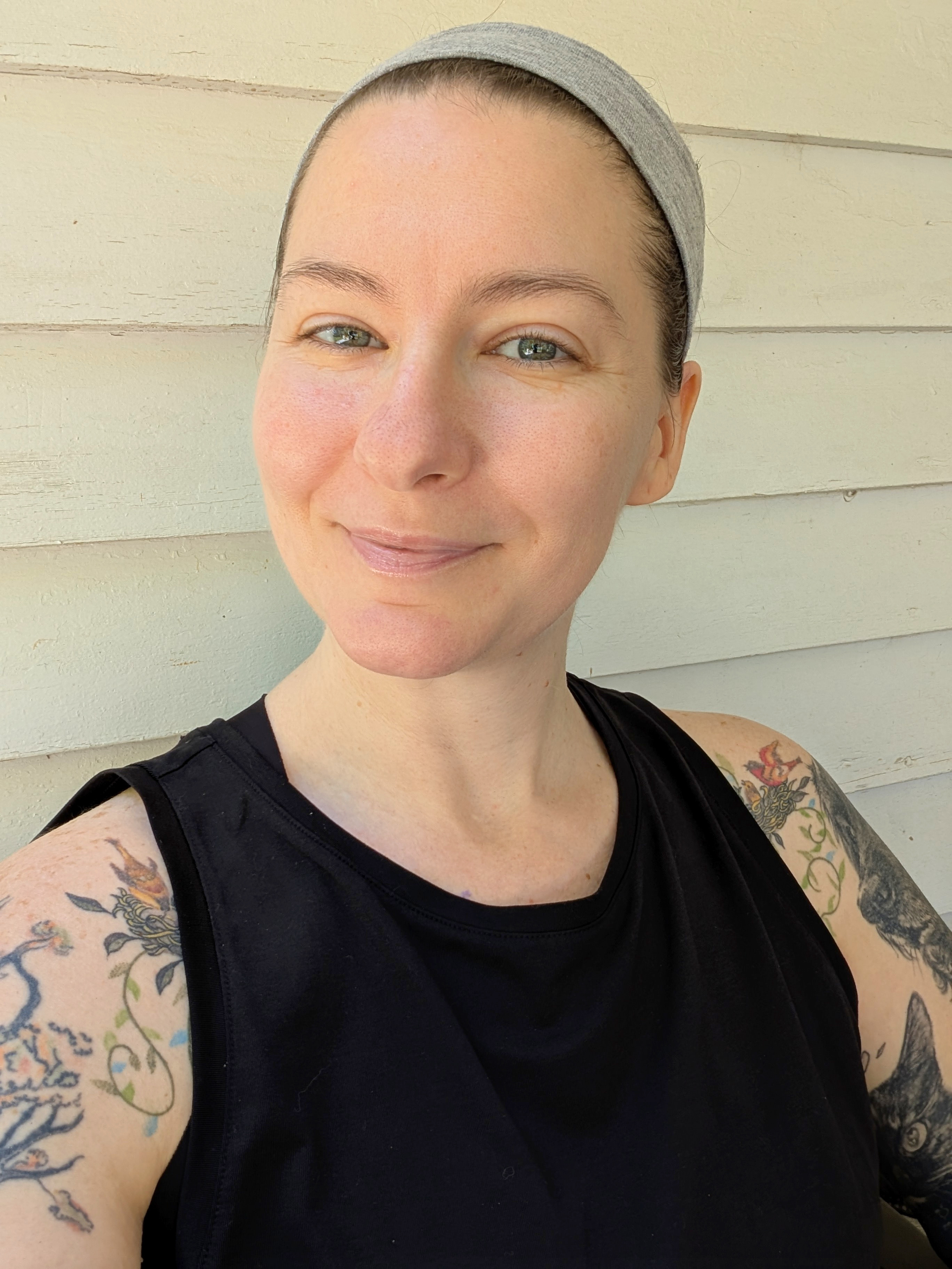 A person with hair pulled back wearing a gray headband and a black sleeveless shirt, smiling, with tattoos, standing in front of a light beige wall.