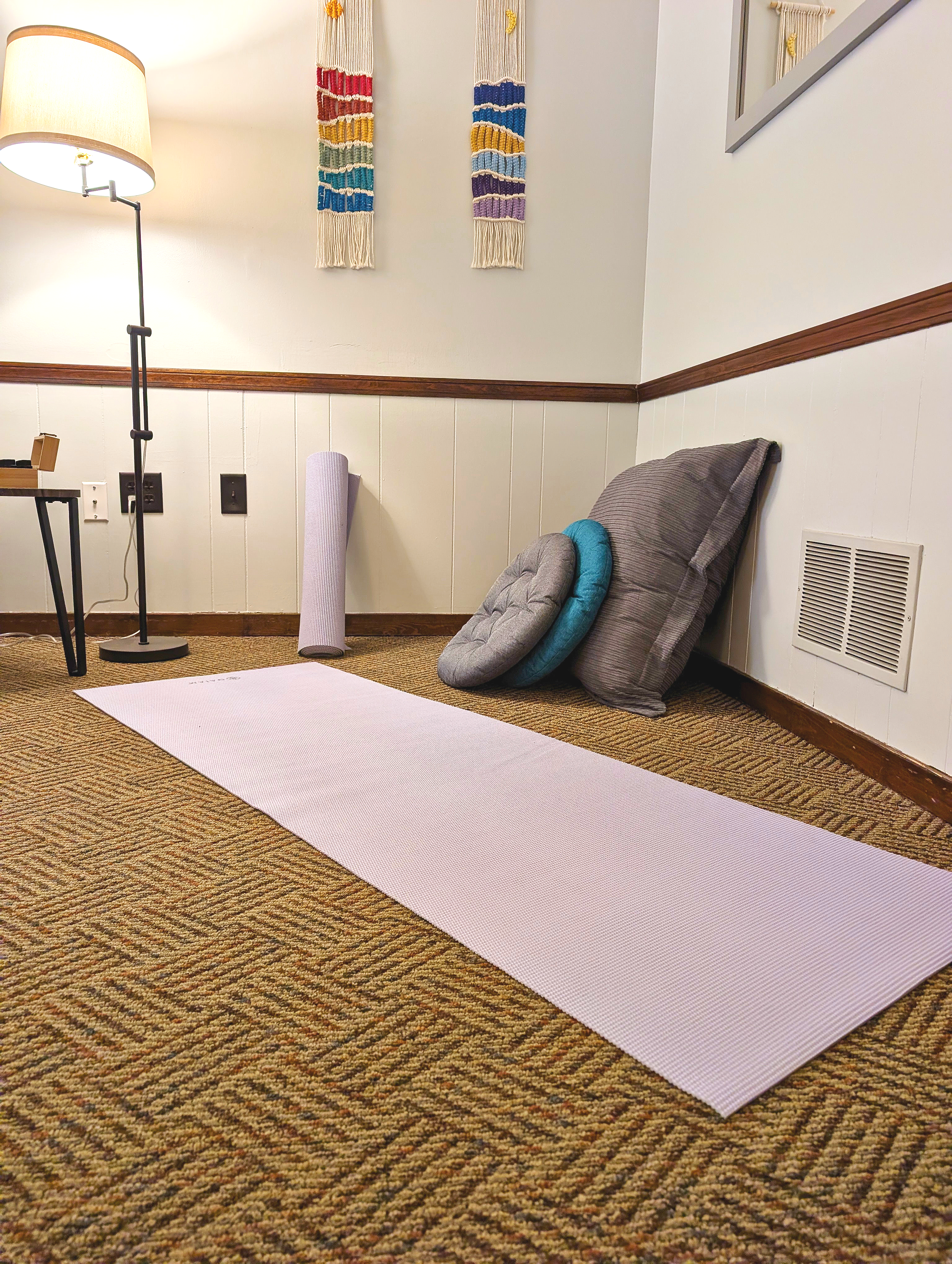 Indoor scene with a lilac yoga mat on a multicolored carpet, floor cushions and large pillow, a tall standing lamp, and wall decorations. The walls are partly white with a wooden chair rail and some woven wall art.