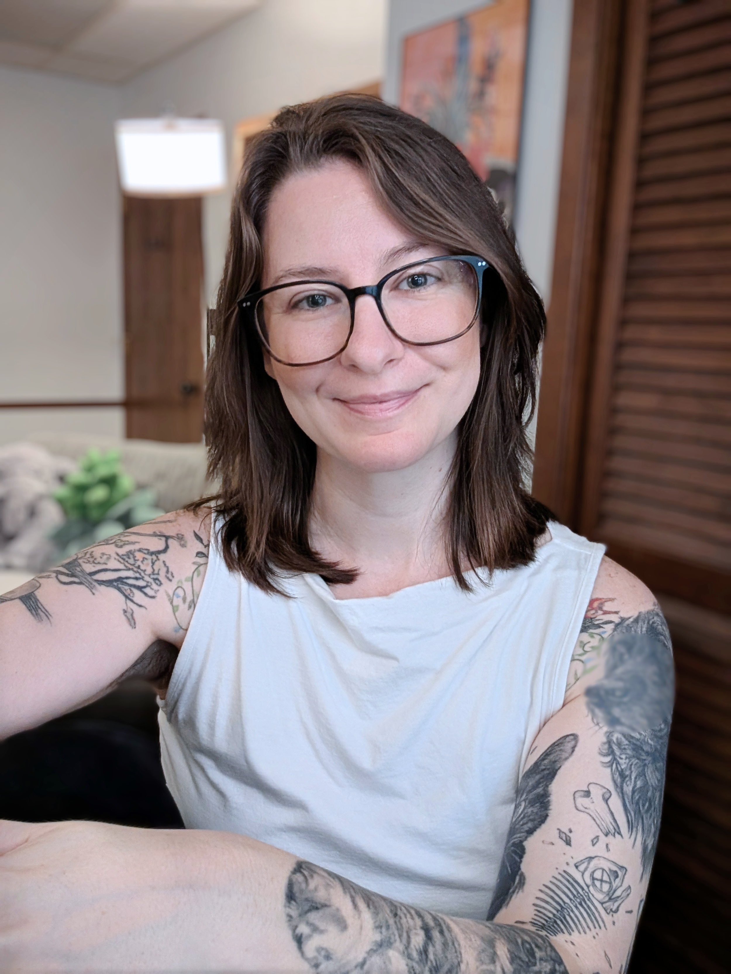 A person with shoulder-length brown hair, glasses, and tattoos is smiling while sitting in a therapy office.