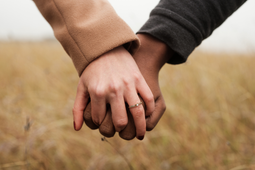 Close-up of two people holding hands, one wearing a ring, with a blurred grassy field in the background.