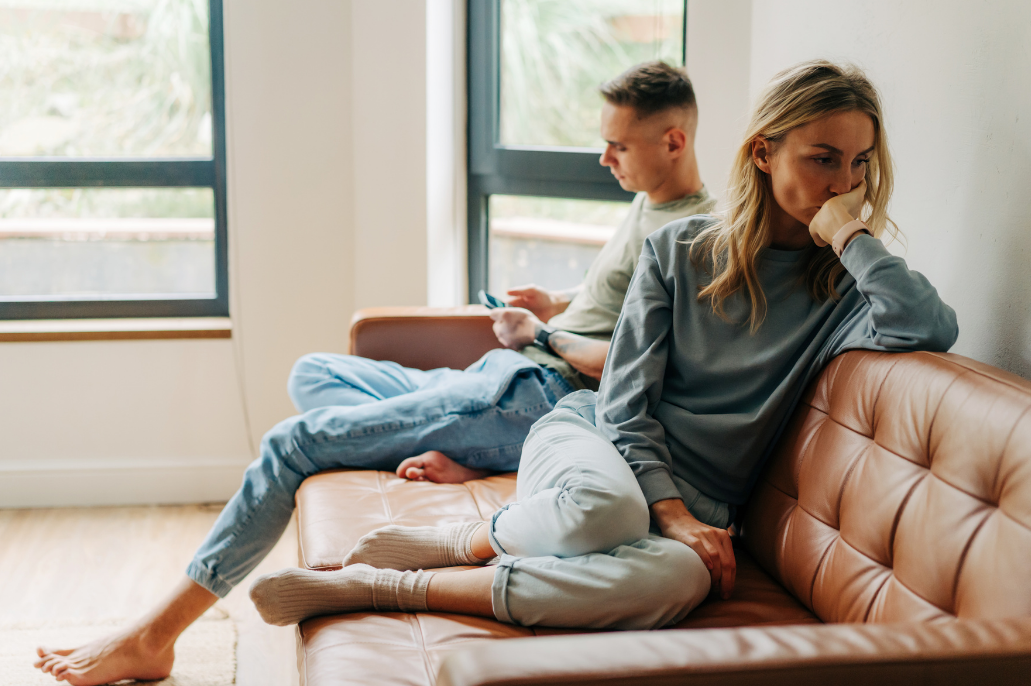 Couple sitting on a sofa, looking distant and thoughtful, with large windows in the background.