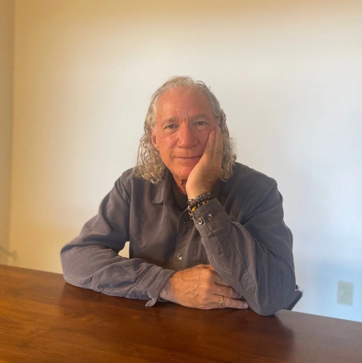 Ben Cohen, Ph.D. who has curly hair and a thoughtful expression, resting his head on his hand, wearing a gray shirt and sitting at his wooden desk.