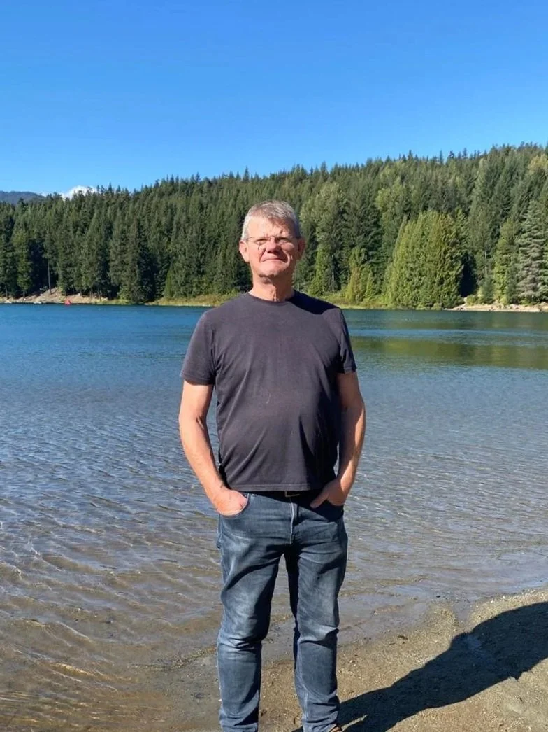 A man standing by a lake with a forested hillside in the background under a clear blue sky.