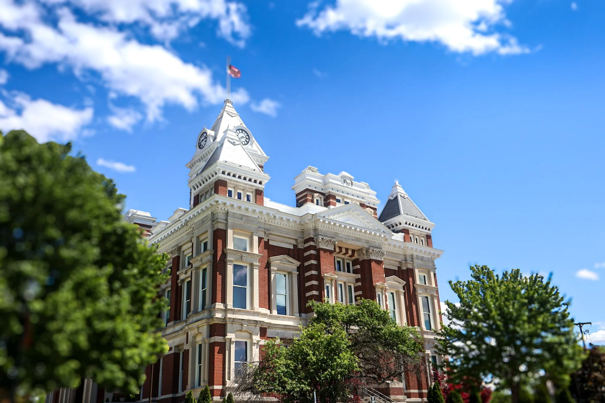 Historic red brick building with white accents, multiple tiers, and clock towers under a blue sky with scattered clouds, partially obscured by green trees.