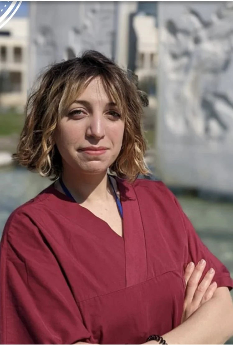 A woman with shoulder-length curly brown hair, wearing a maroon shirt, standing outdoors with her arms crossed near a metallic sculpture and a modern building in the background.