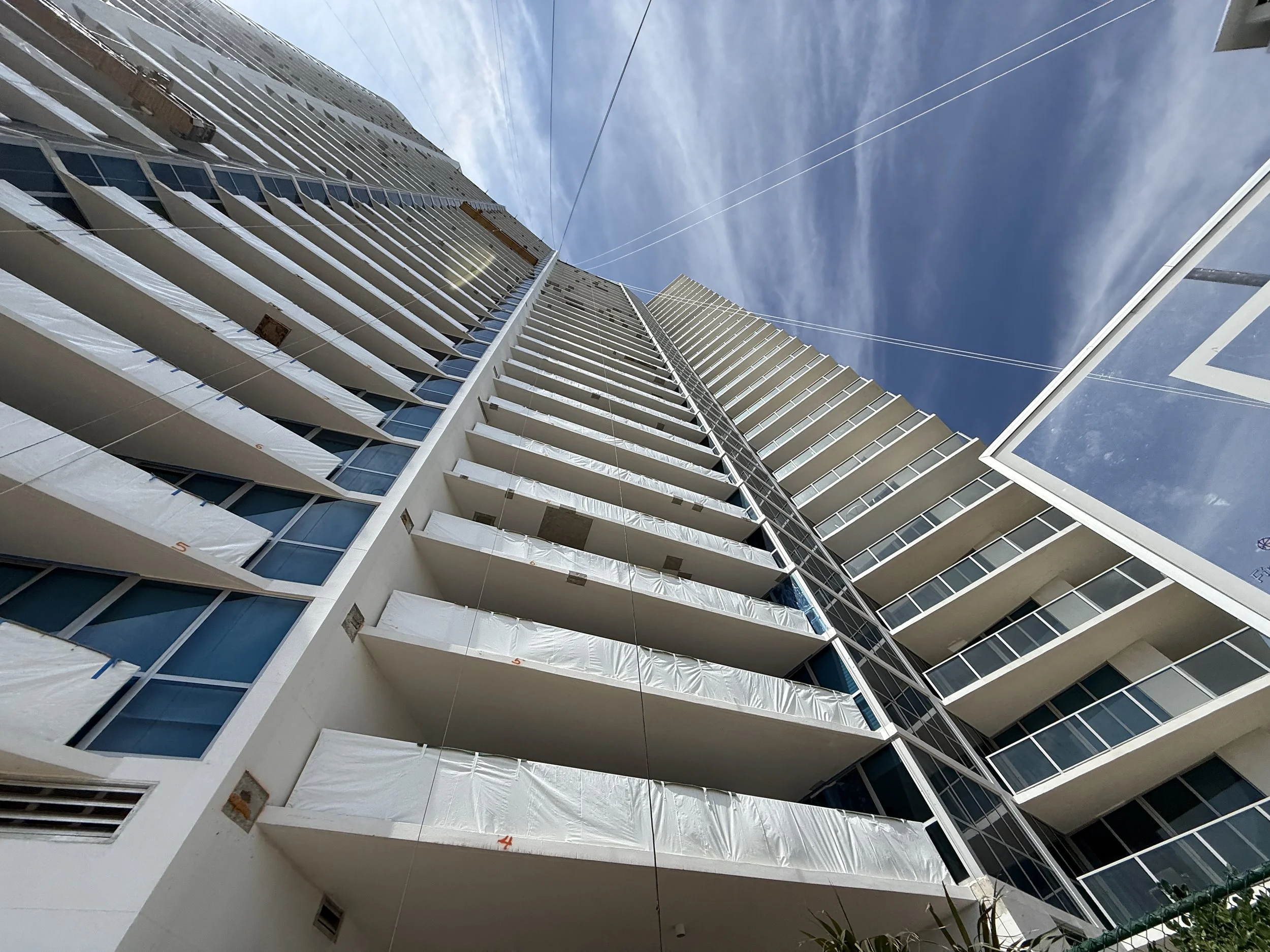 Low-angle view of a modern high-rise building with multiple balconies and blue tinted windows, against a bright blue sky with wispy clouds.