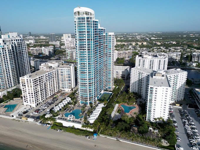 A high-rise modern hotel or apartment building with a curved design, surrounded by other buildings, swimming pools, and parking lots in an urban area.