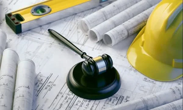 Construction desk with blueprints, a yellow safety helmet, a gavel, a spirit level, a measuring tape, and rolled blueprints.