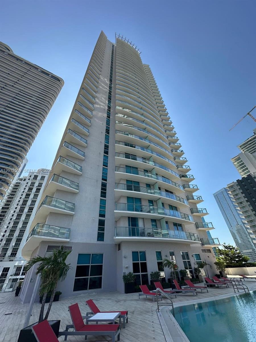 A tall modern high-rise building with balconies on each floor, situated next to other skyscrapers, with a pool area featuring lounge chairs and palm trees in the foreground.