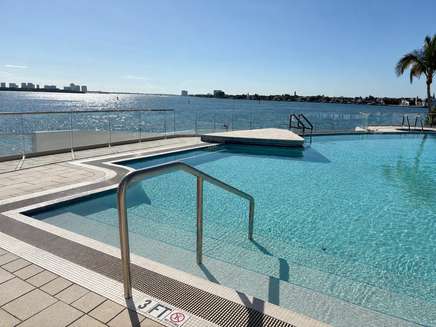 A swimming pool with clear blue water, metal handrails, and a small ledge, next to a body of water, under a sunny sky with a city skyline in the background.