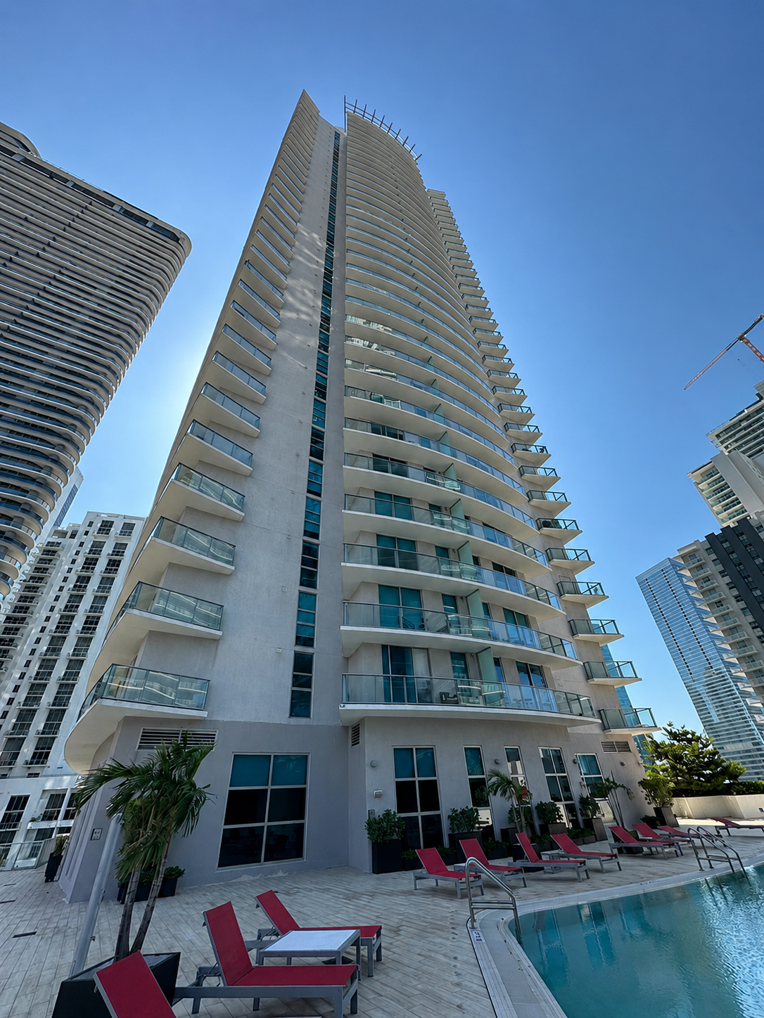 A modern high-rise building with multiple balconies, situated next to other tall buildings, features a rooftop pool area with lounge chairs, potted plants, and a clear blue sky in the background.