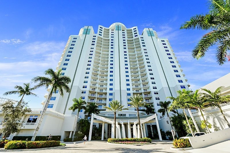 Tall white beachfront hotel with curved balconies, surrounded by palm trees under a blue sky.
