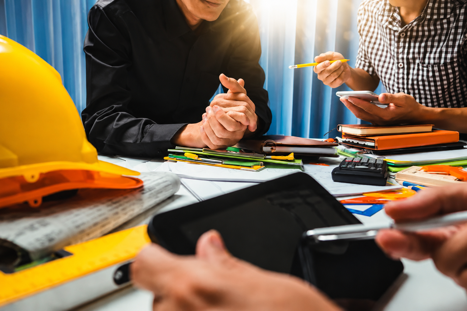 Group of engineers working together at a table covered with safety helmets, notebooks, pens, calculators, and smartphones during a meeting.