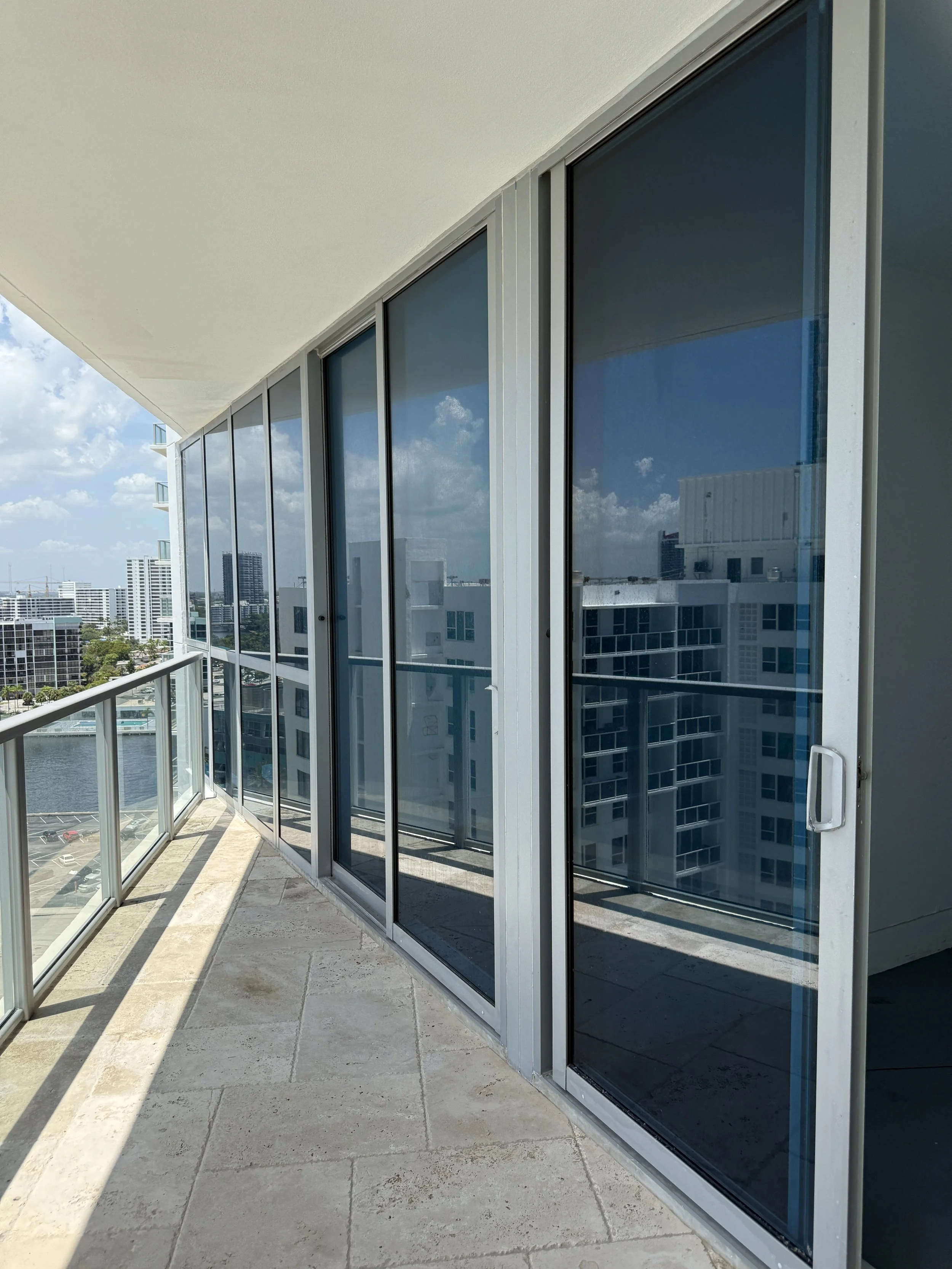 Balcony with glass doors and railing, overlooking a city skyline with modern high-rise buildings and water body, under a partly cloudy sky.