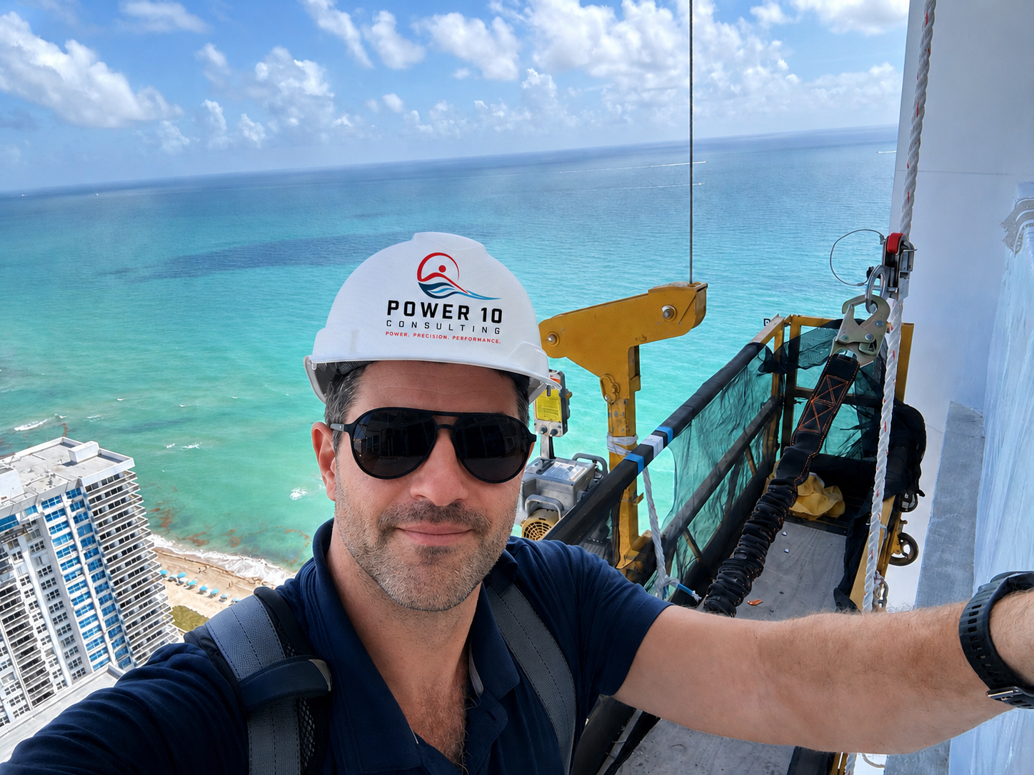 A man in sunglasses and a white hard hat taking a selfie from a high-rise building rooftop. The background shows a view of the ocean with a beach and a tall building. Construction equipment and safety harnesses are visible on the rooftop.
