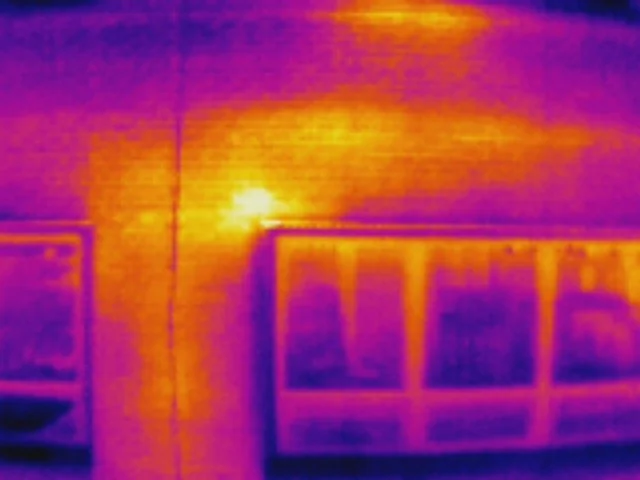 Infrared thermal image of a building exterior showing heat patterns, including a bright spot on the wall indicating a heat source.