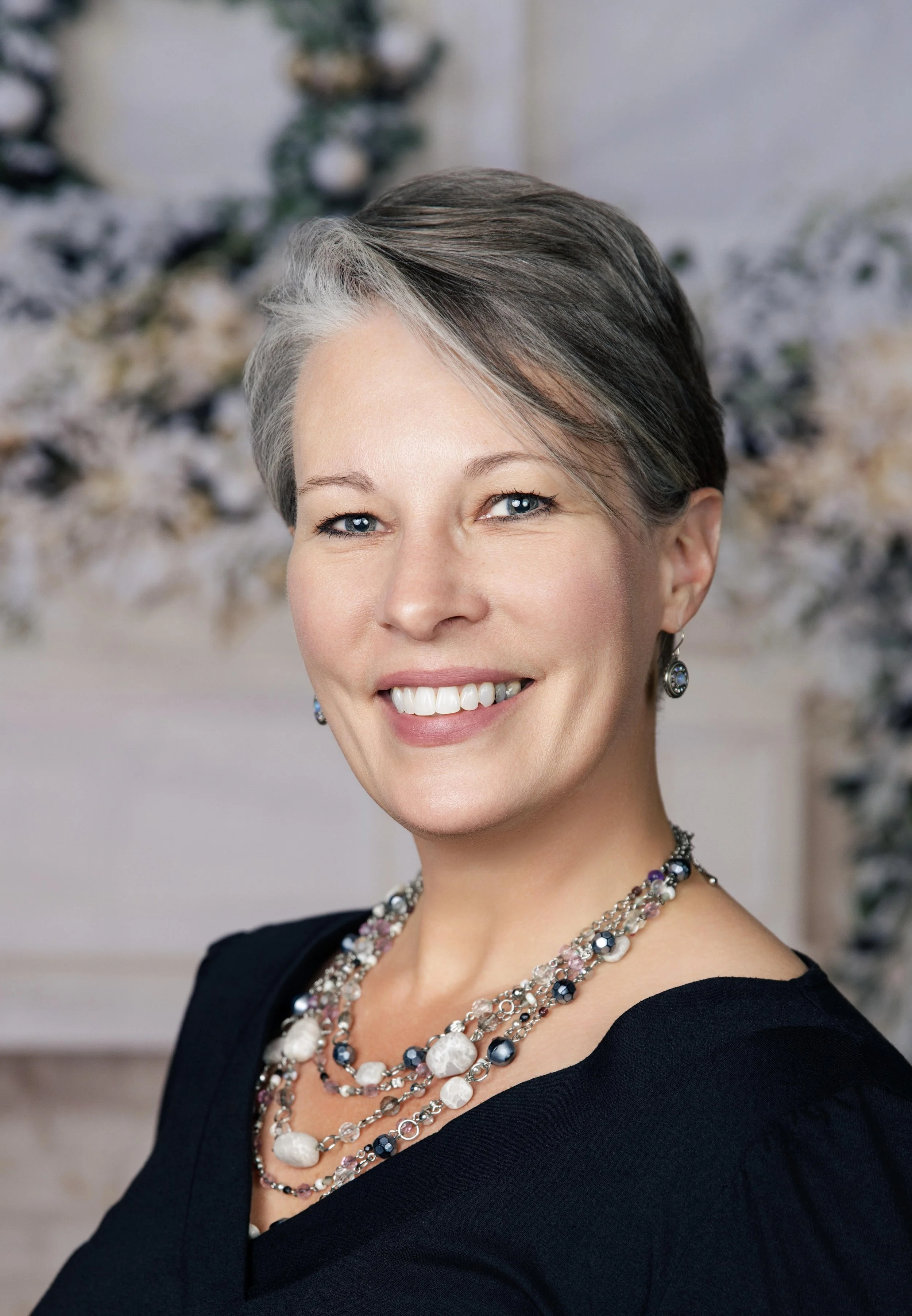 A woman with short gray and brown hair, blue eyes, wearing a black dress, colorful jewelry including necklaces, earrings, and a choker, smiling in front of a blurred floral background.