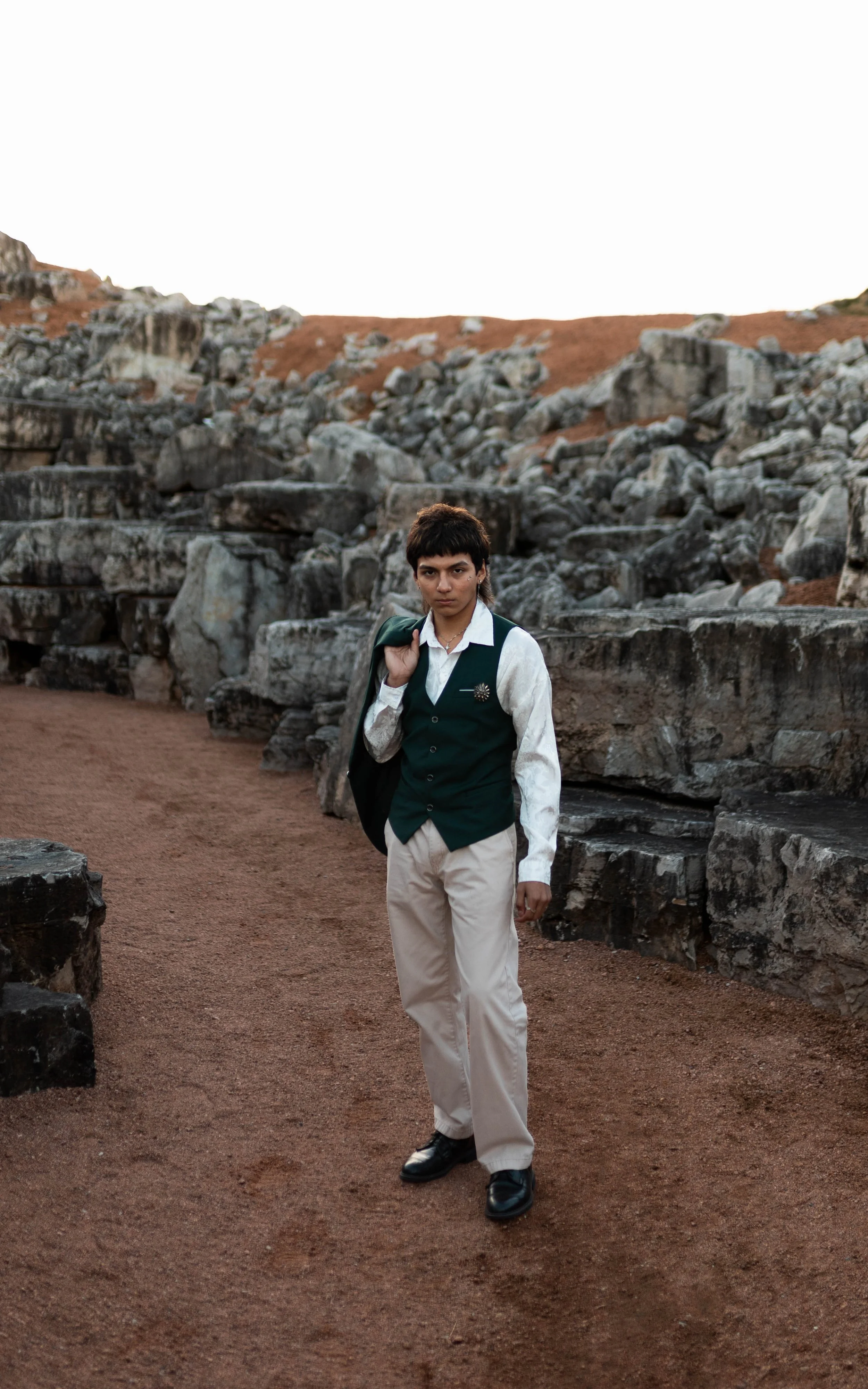 Young person with short dark hair standing on a dirt path surrounded by rocks and a rocky hillside, wearing a green vest over a white shirt and beige pants, holding a jacket over their shoulder, with a serious expression.