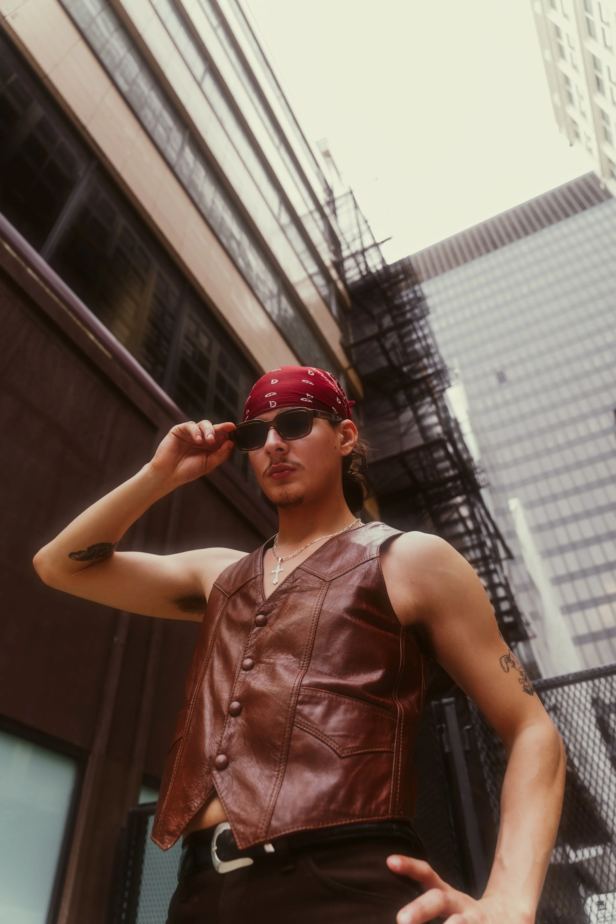 A man wearing sunglasses and a red bandana adjusts his glasses while standing outdoors in an urban setting with tall buildings and fire escapes.