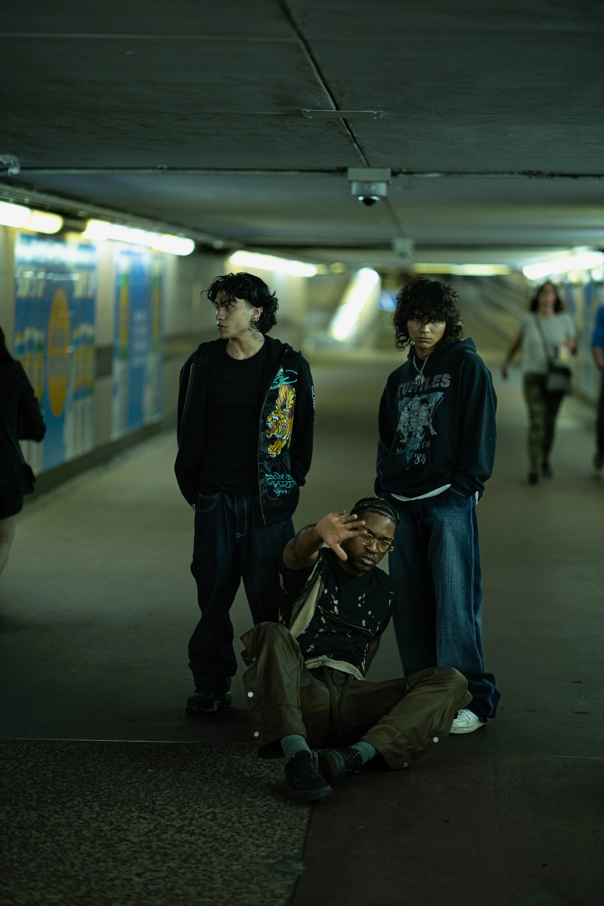 Three young men in casual clothing are in an underground tunnel or subway station. Two are standing, and one is sitting or crouching on the ground, with the seated man holding his hand up as if waving or blocking the camera. The background shows blurred lights and advertisements.