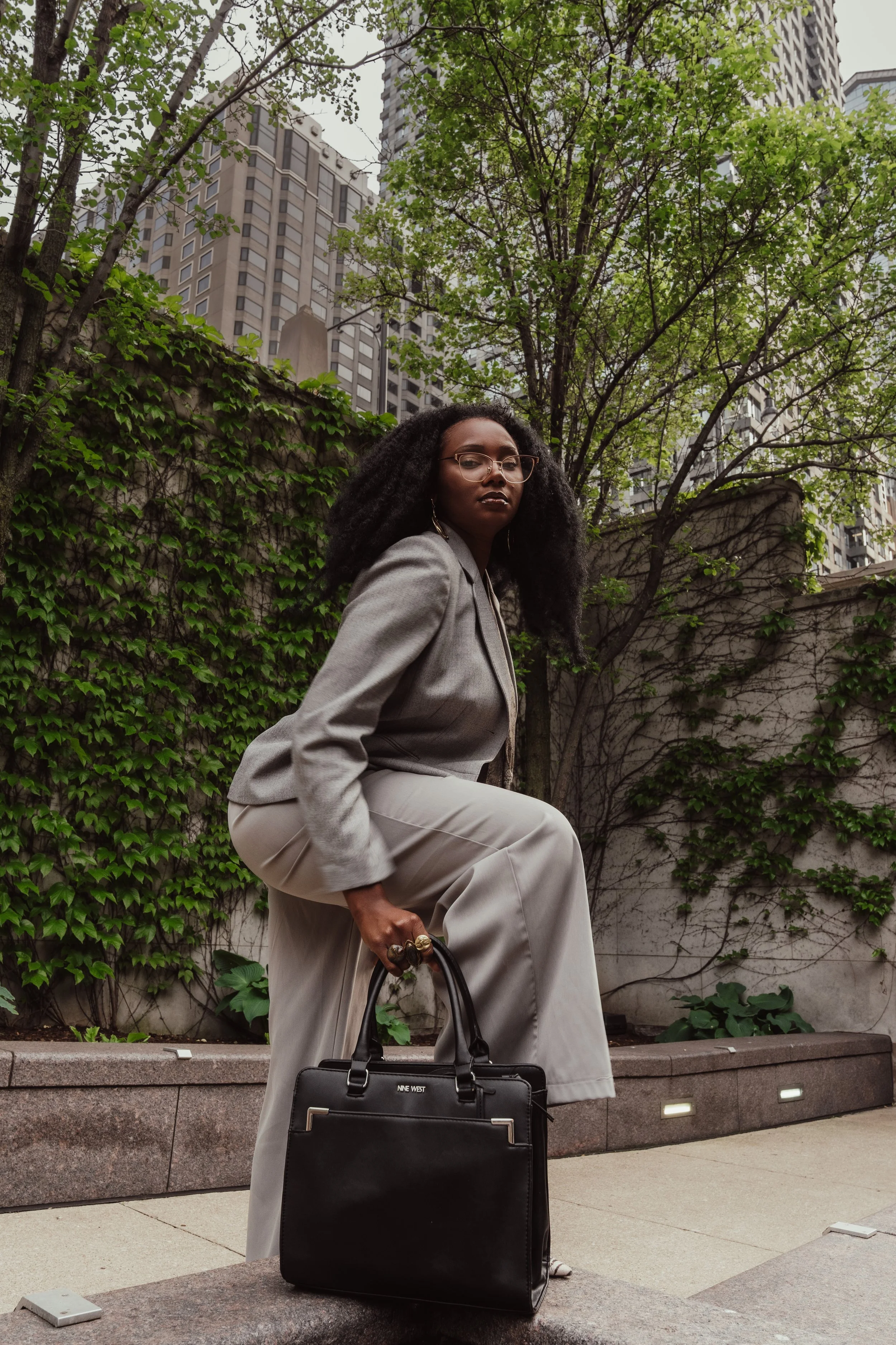 A woman with curly black hair, wearing glasses and a gray blazer, posing outdoors in front of a brick wall covered with green ivy and tall buildings in the background. She is holding a black tote bag and has a serious expression.