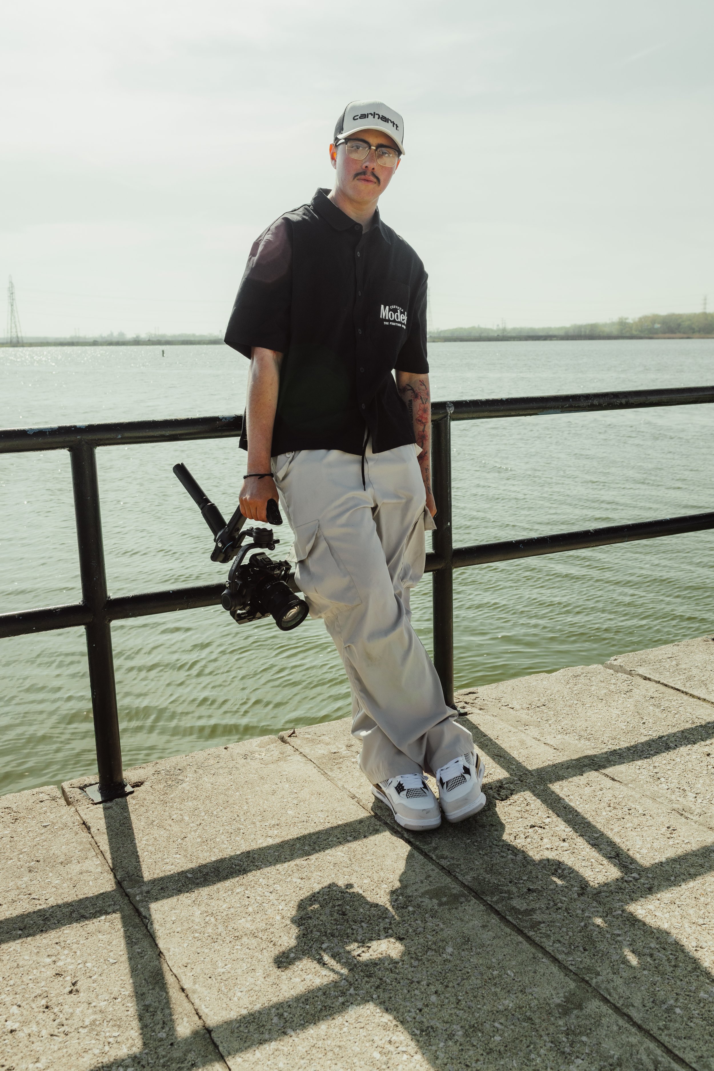 Young man with glasses, a mustache, wearing a black shirt with a logo, beige pants, white sneakers, and a gray cap, standing by a railing by a body of water, holding a camera, with trees and power lines in the background on a cloudy day.