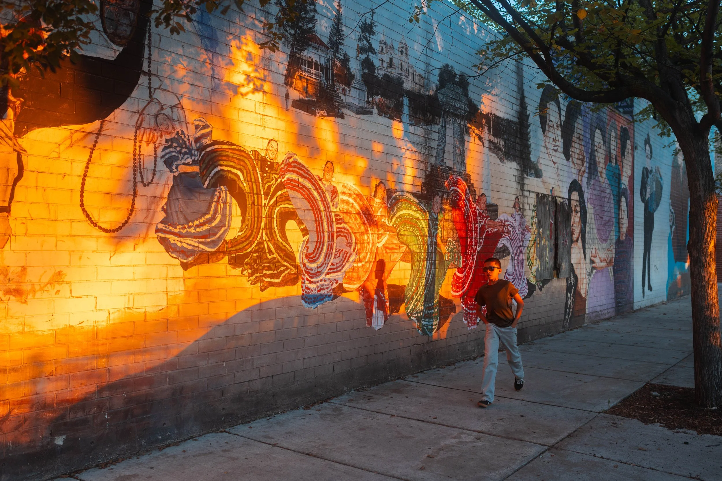 A mural of dancing women painted on a brick wall, illuminated by streetlight, with a young man walking past on the sidewalk and a tree partially covering the mural.