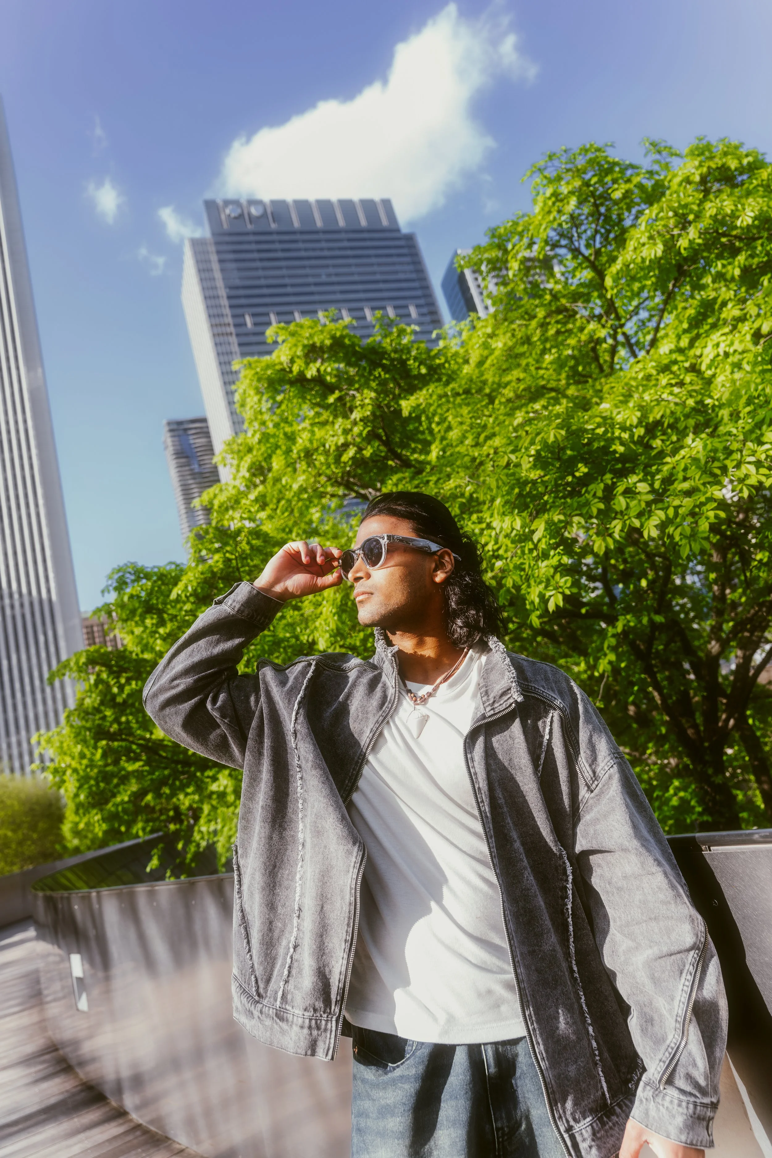 A man with long dark hair wearing sunglasses, a white t-shirt, and a black denim jacket, standing outdoors with green trees and tall buildings in the background.